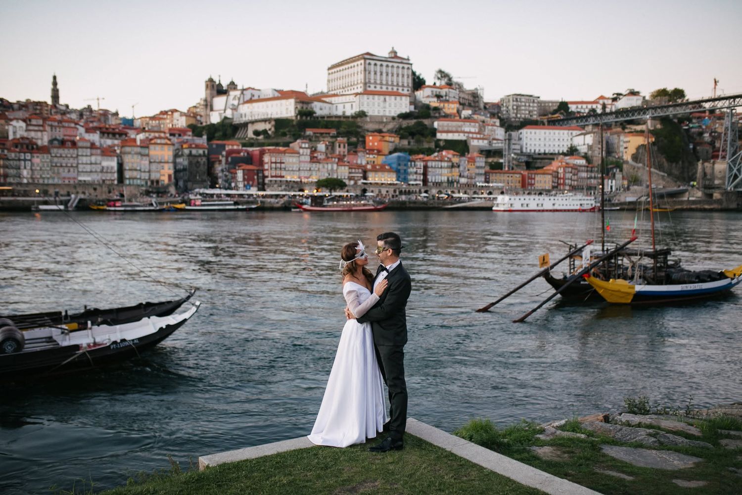 Couple embracing in wedding attire by the Douro River with Porto's colorful historic skyline and boats in the background.