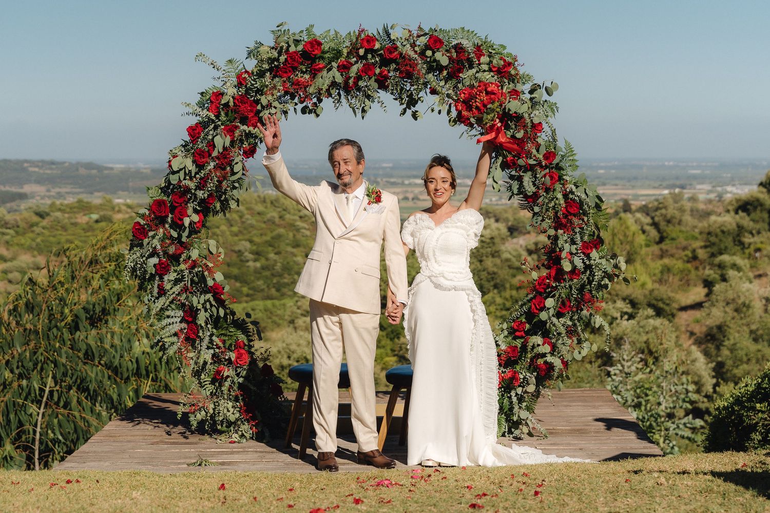 Couple poses under a stunning red rose arch at their outdoor hilltop wedding ceremony.