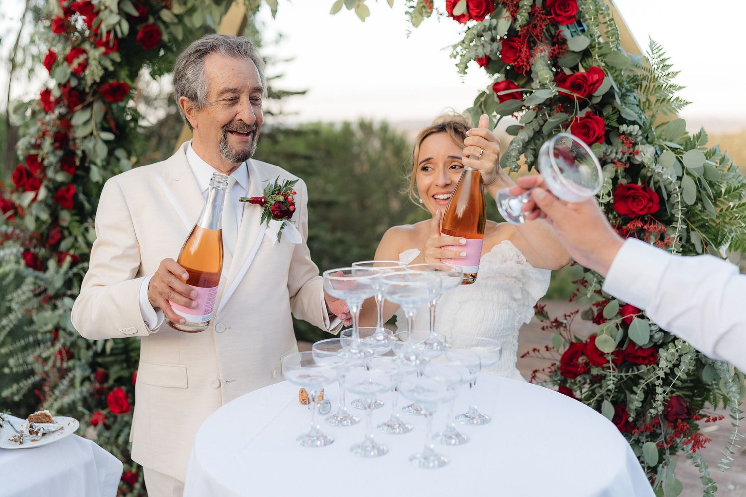Wedding party pouring champagne for celebration toast under red rose arch.