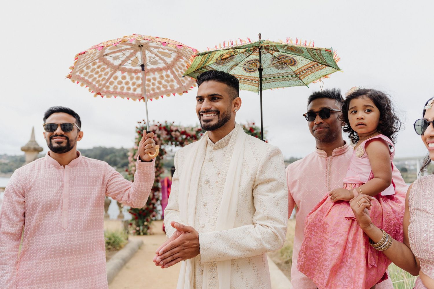 Wedding party members in pink and white attire celebrating outdoors under umbrellas.