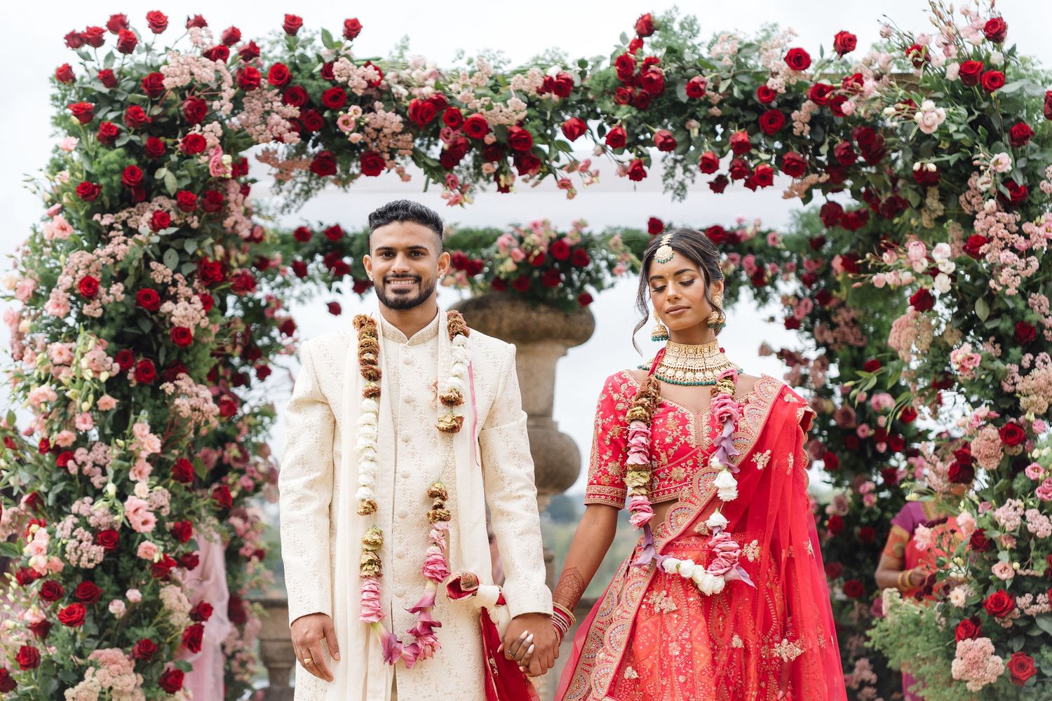 Wedding celebration under red rose archway with guests in traditional Indian formal wear.