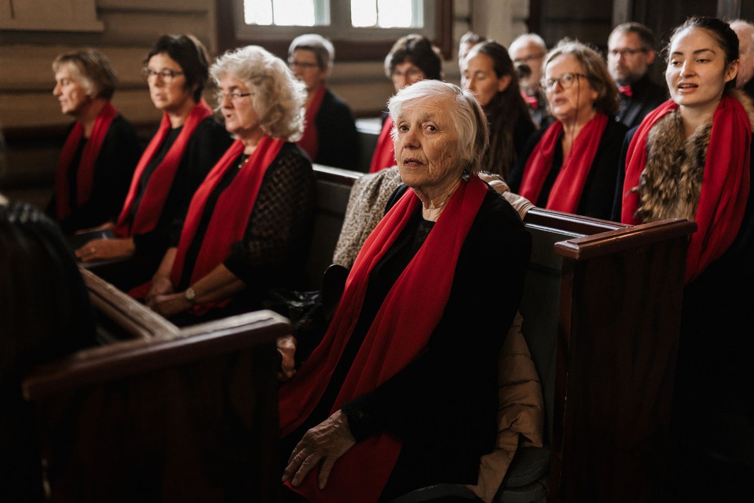 Members of a choir in red scarves stand in rows at a formal indoor gathering.