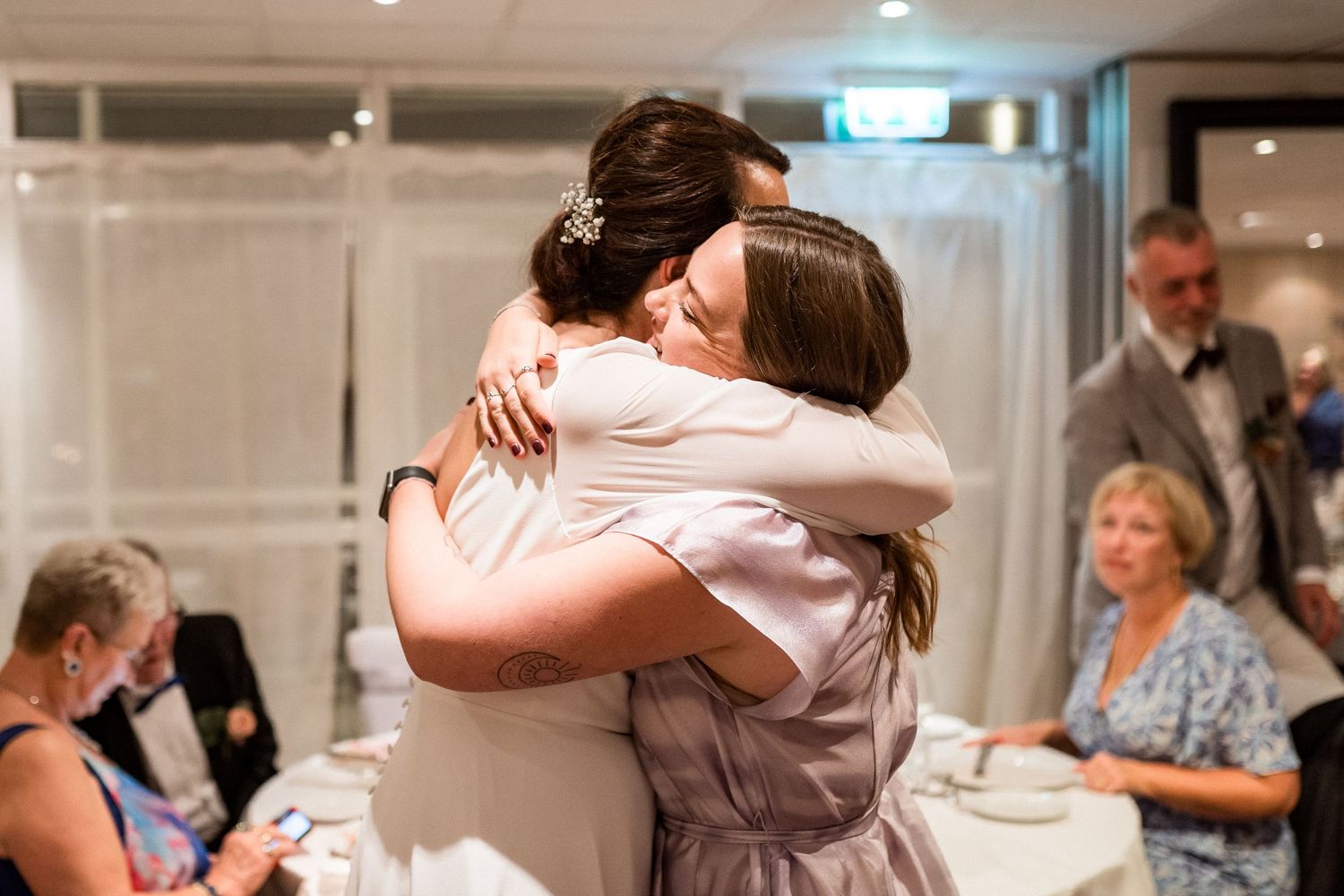 Wedding reception guests sharing emotional embraces in a bright indoor venue.