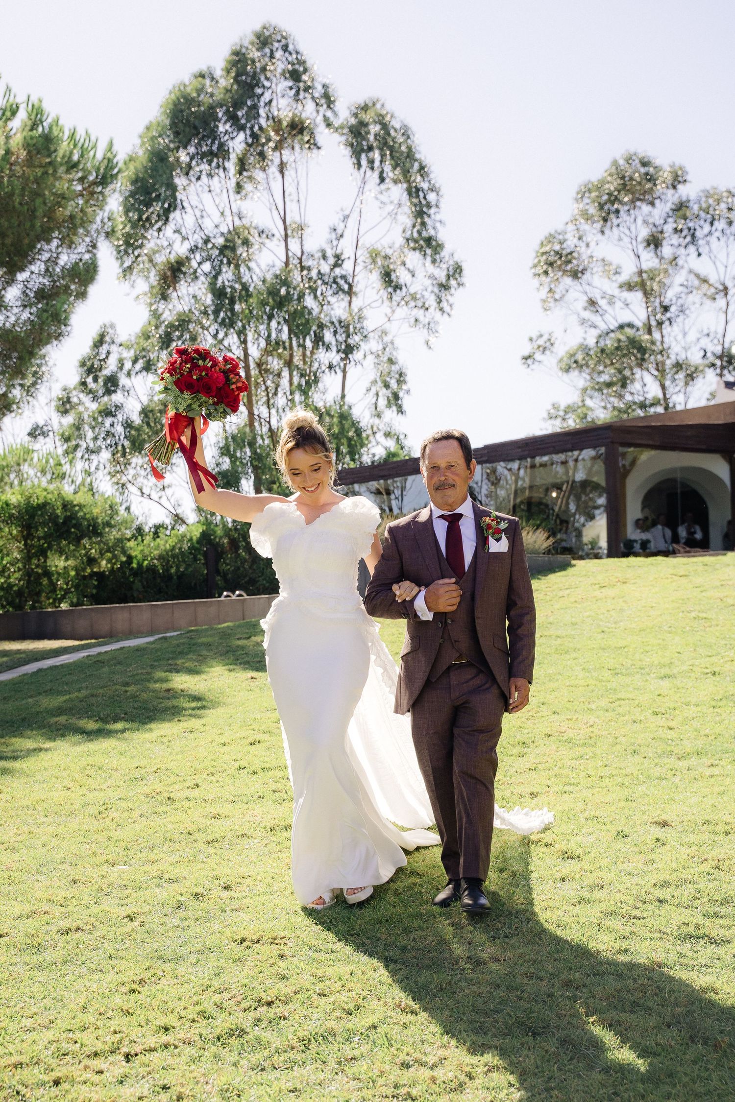 Wedding couple walks hand in hand across sunny hillside with eucalyptus trees.