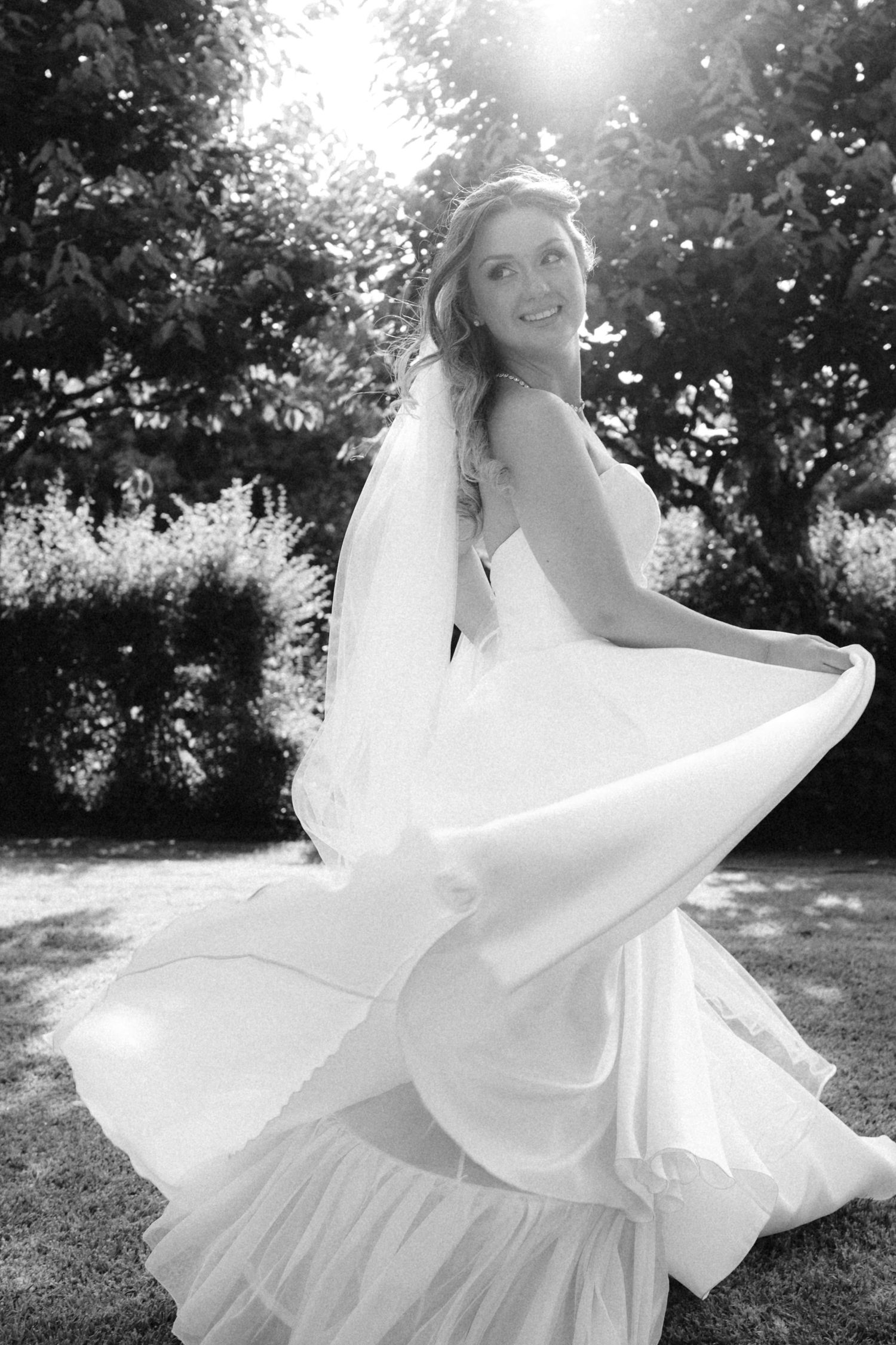 Bride twirling in a flowing white wedding dress outdoors in sunlit garden setting.