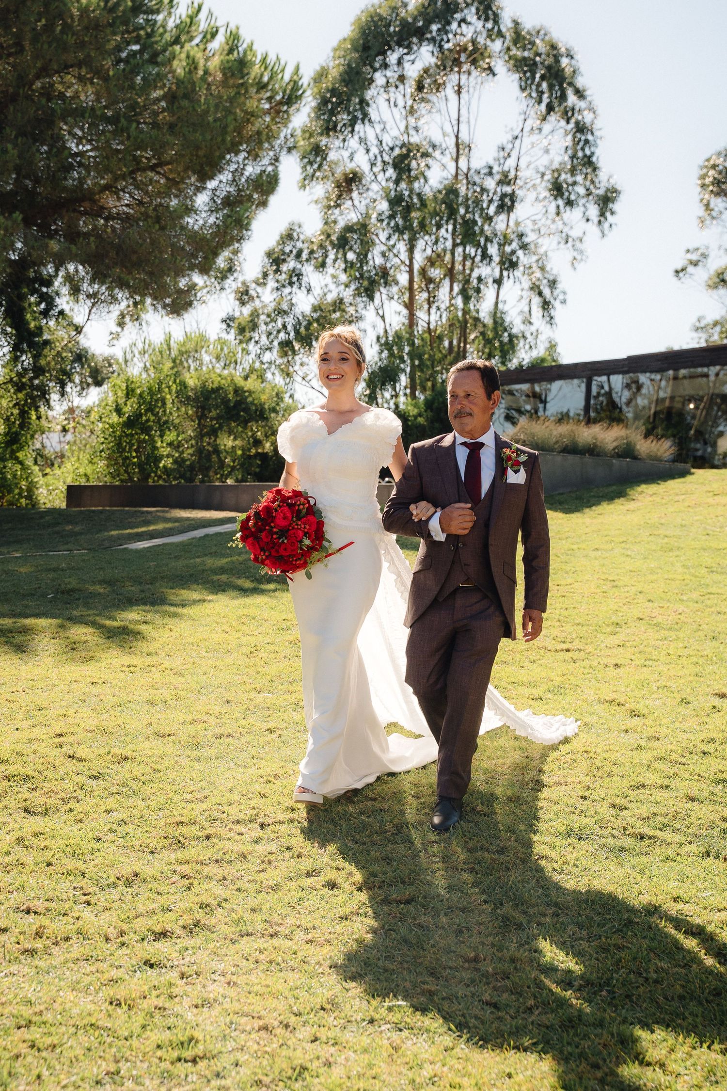 A couple walks together on grass, with bride in white dress holding vibrant red bouquet.