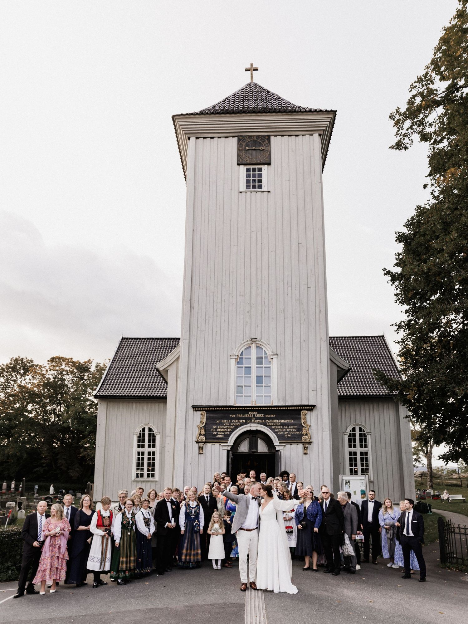 White wooden church exterior with wedding party and guests gathered for group photo.