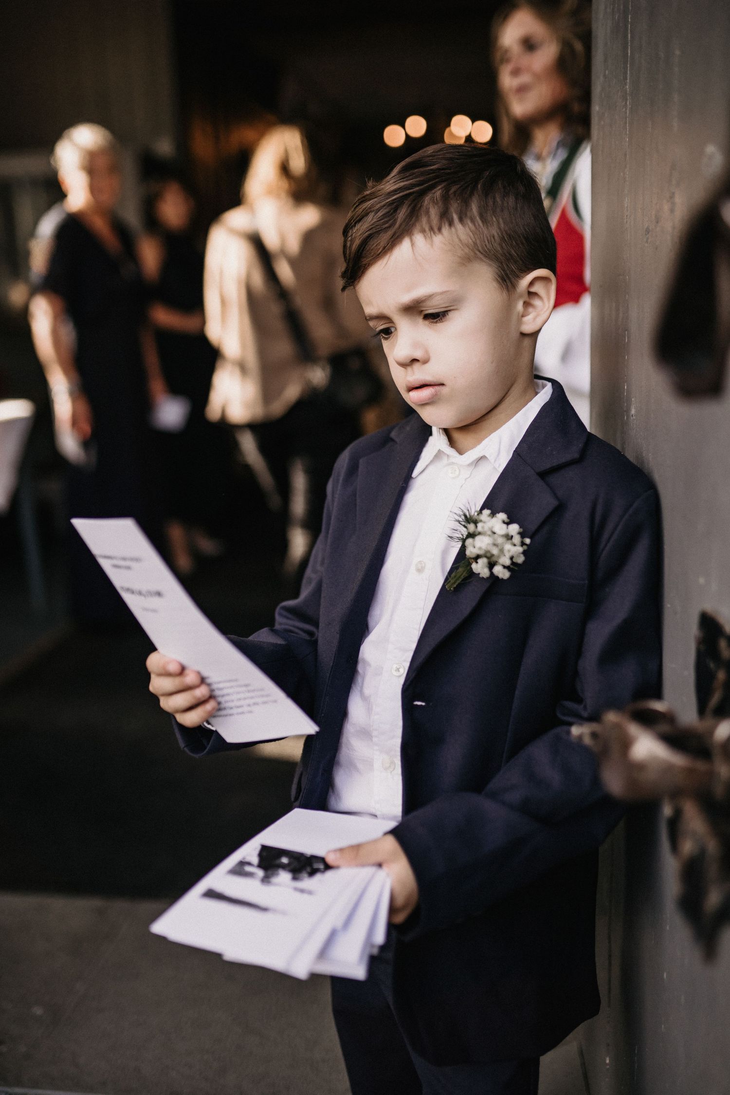 Young boy in a navy suit reads a program during a formal ceremony.