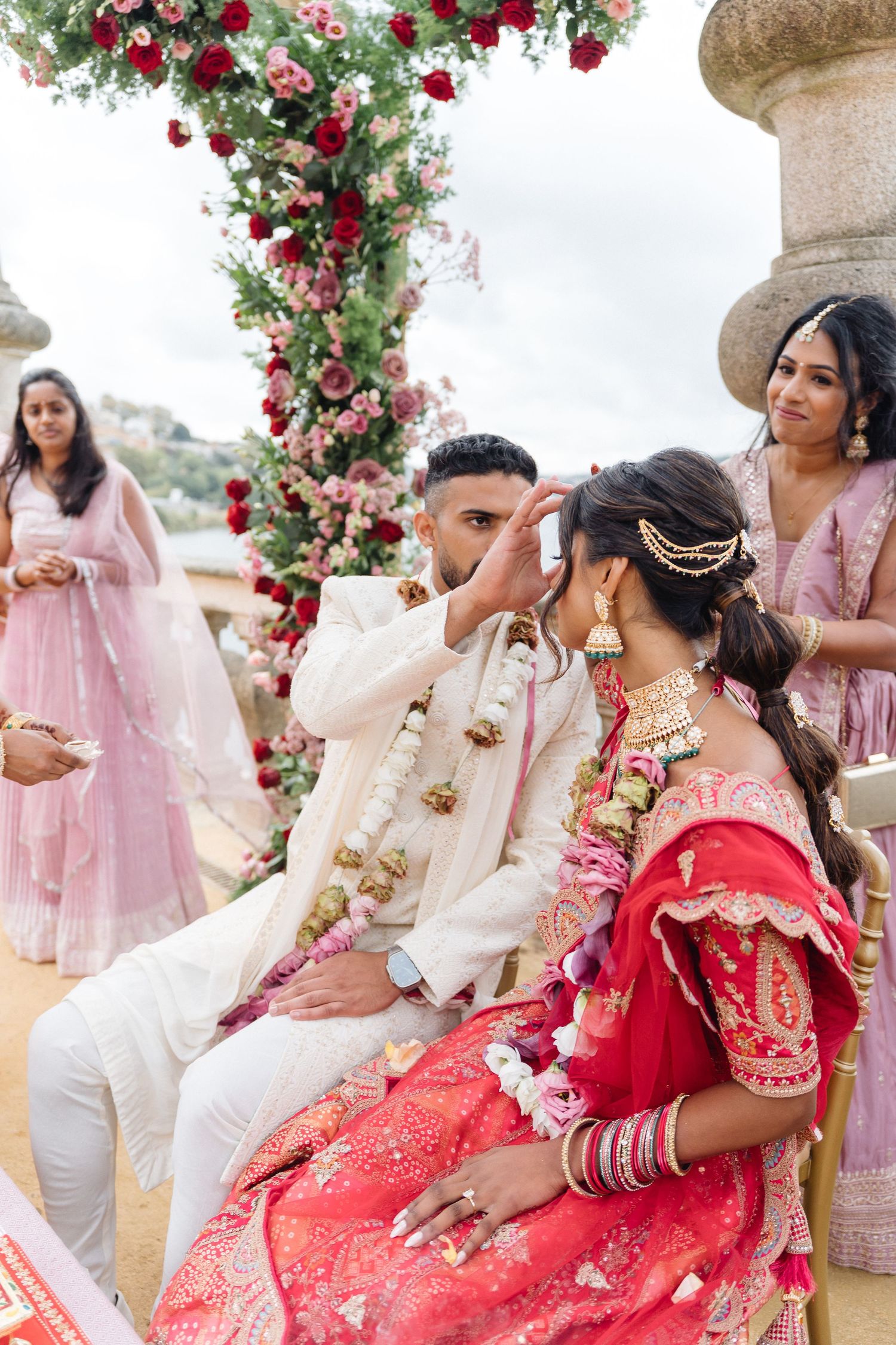 Wedding party members in pink and red attire pose under a floral arch during an outdoor celebration.