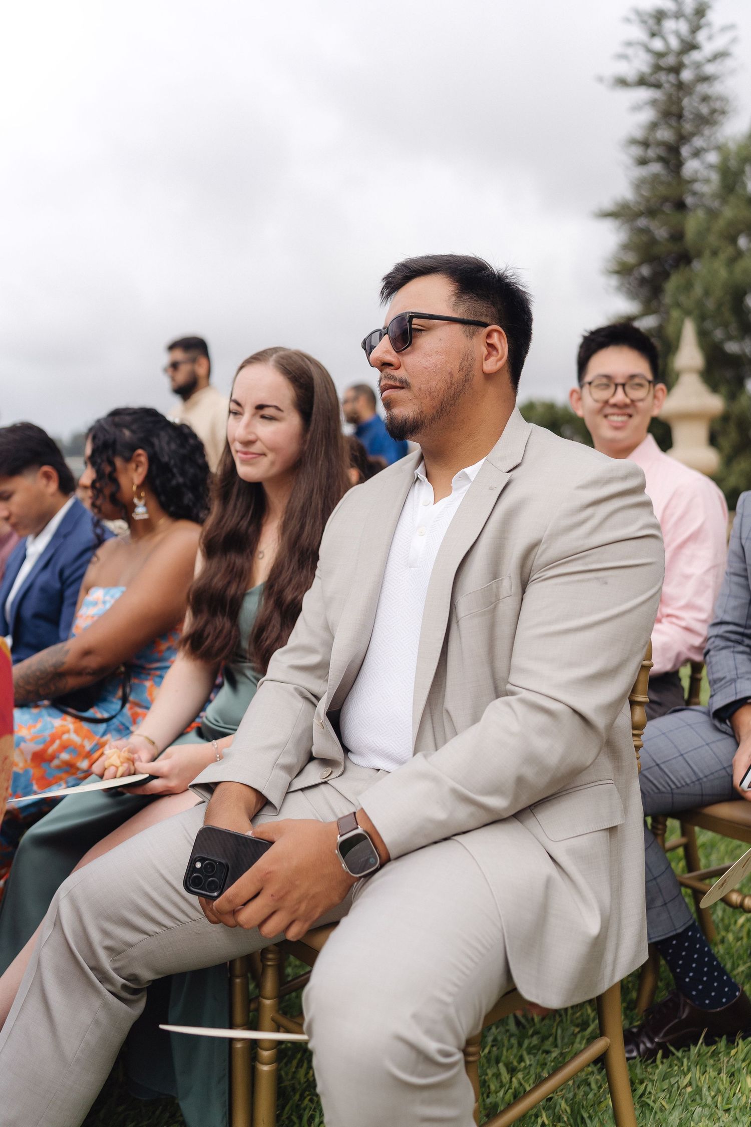 Wedding guests in formal attire seated on wooden chairs during outdoor ceremony.