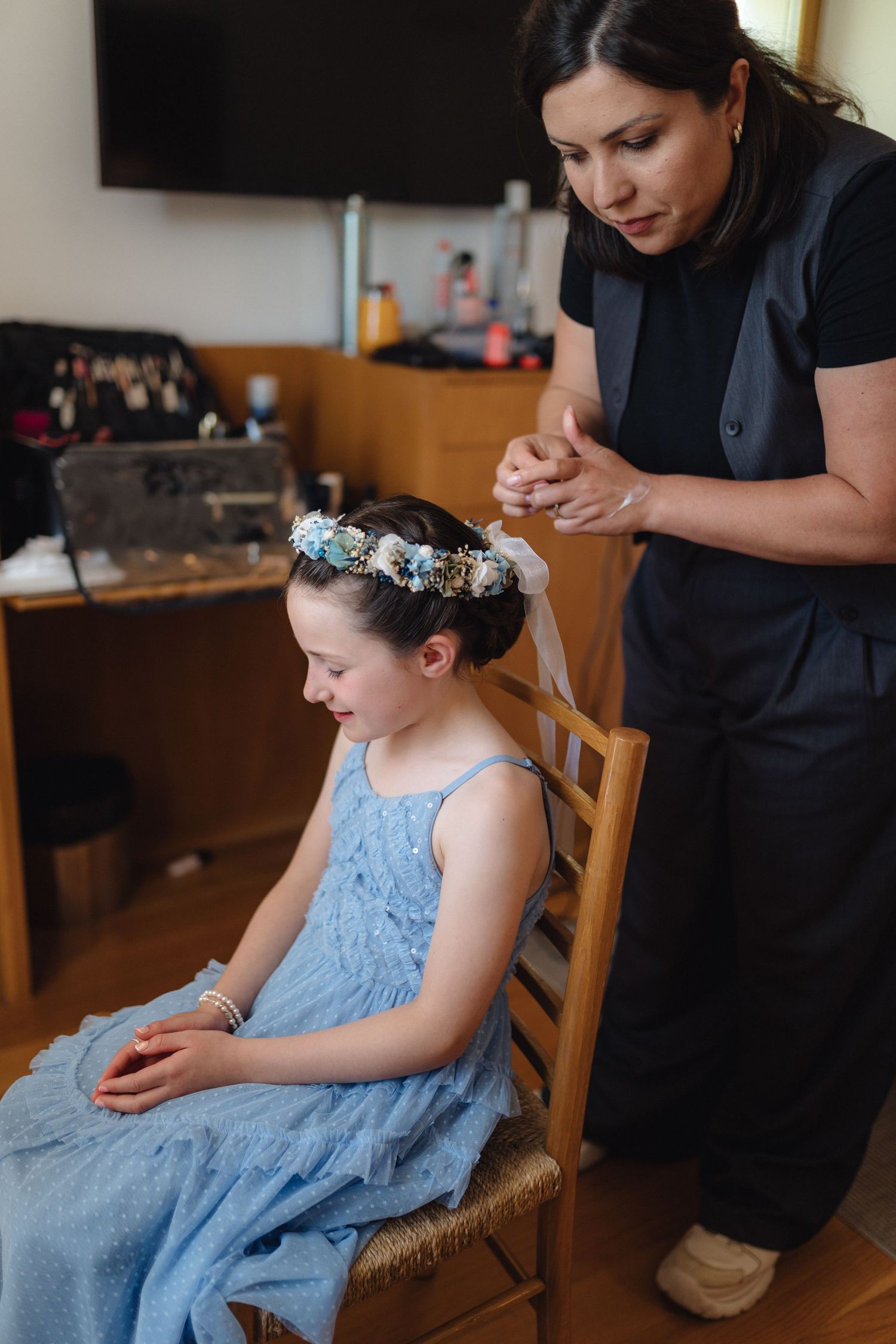 Young person in a blue dress getting their hair styled while seated in a wooden chair.