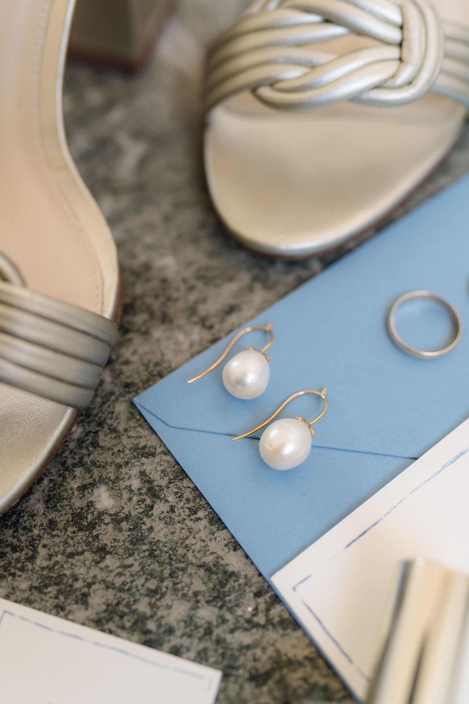 Pearl earrings and elegant bridal shoes arranged with blue stationery on marble surface.