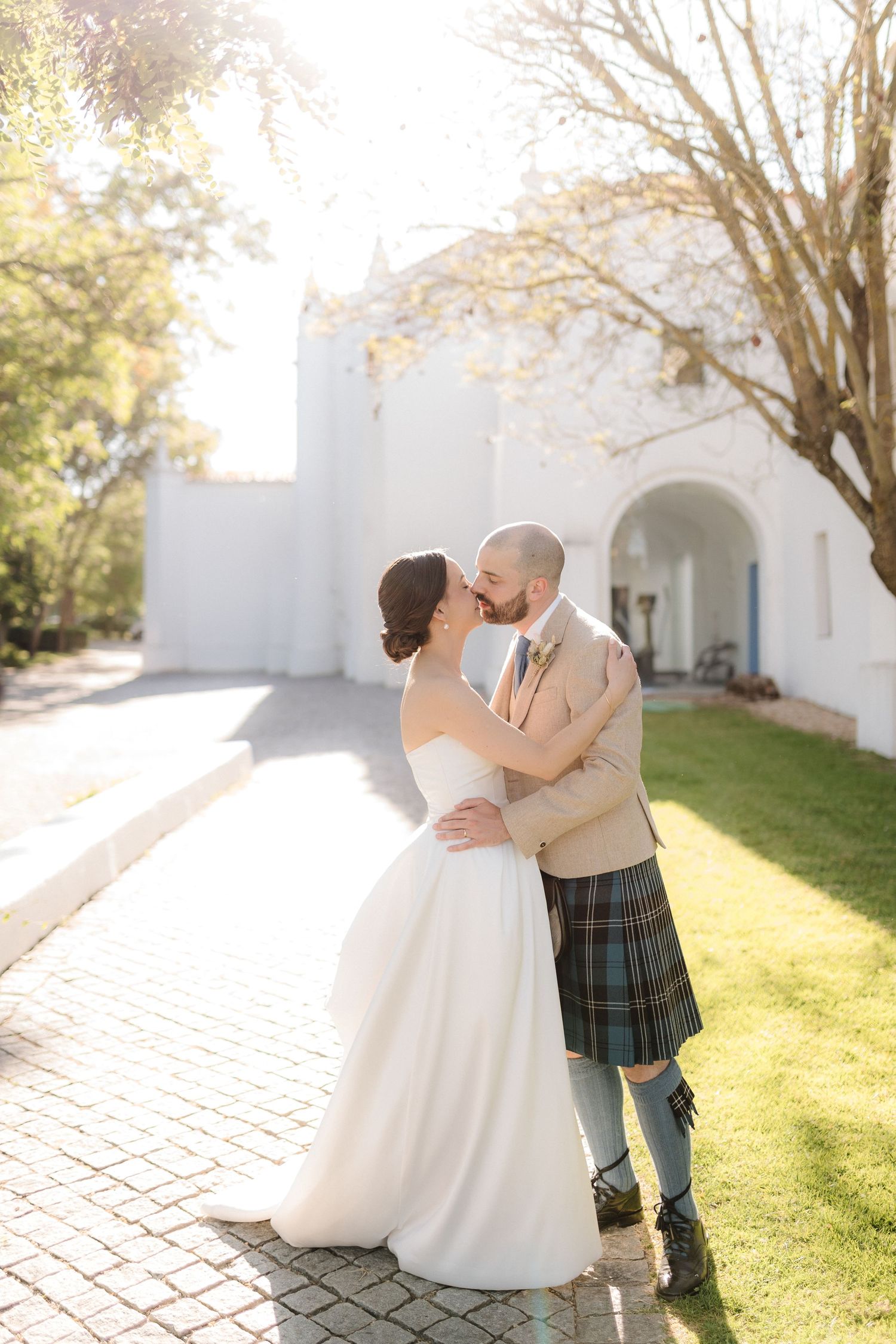 Romantic wedding couple embracing in sunlit garden with white church walls and cobblestone path.