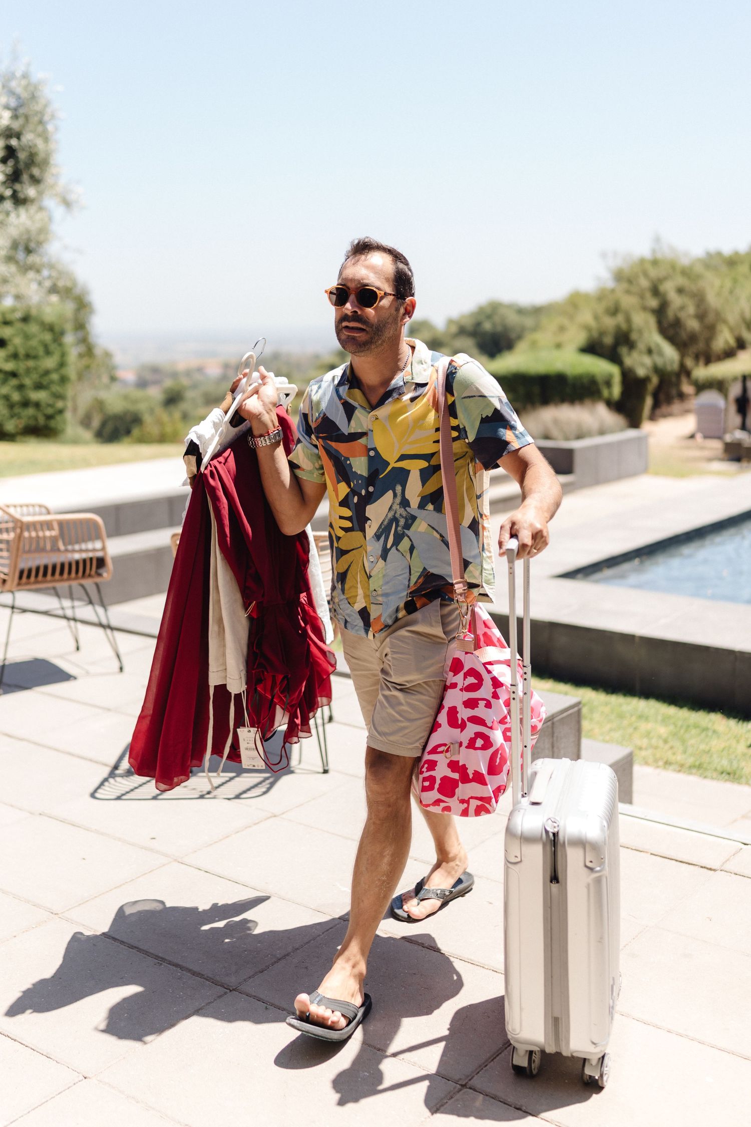 Person carrying luggage and towels on a sunny pool deck with scenic views.