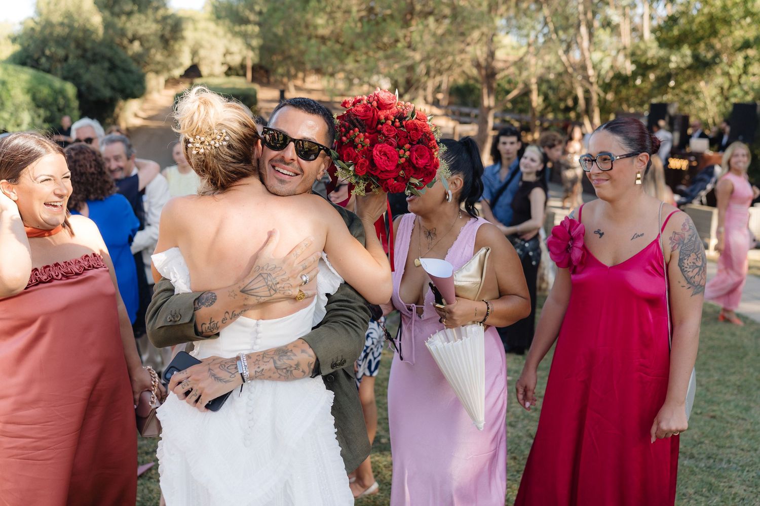 Wedding party members in pink and red dresses share embraces on a grassy lawn.