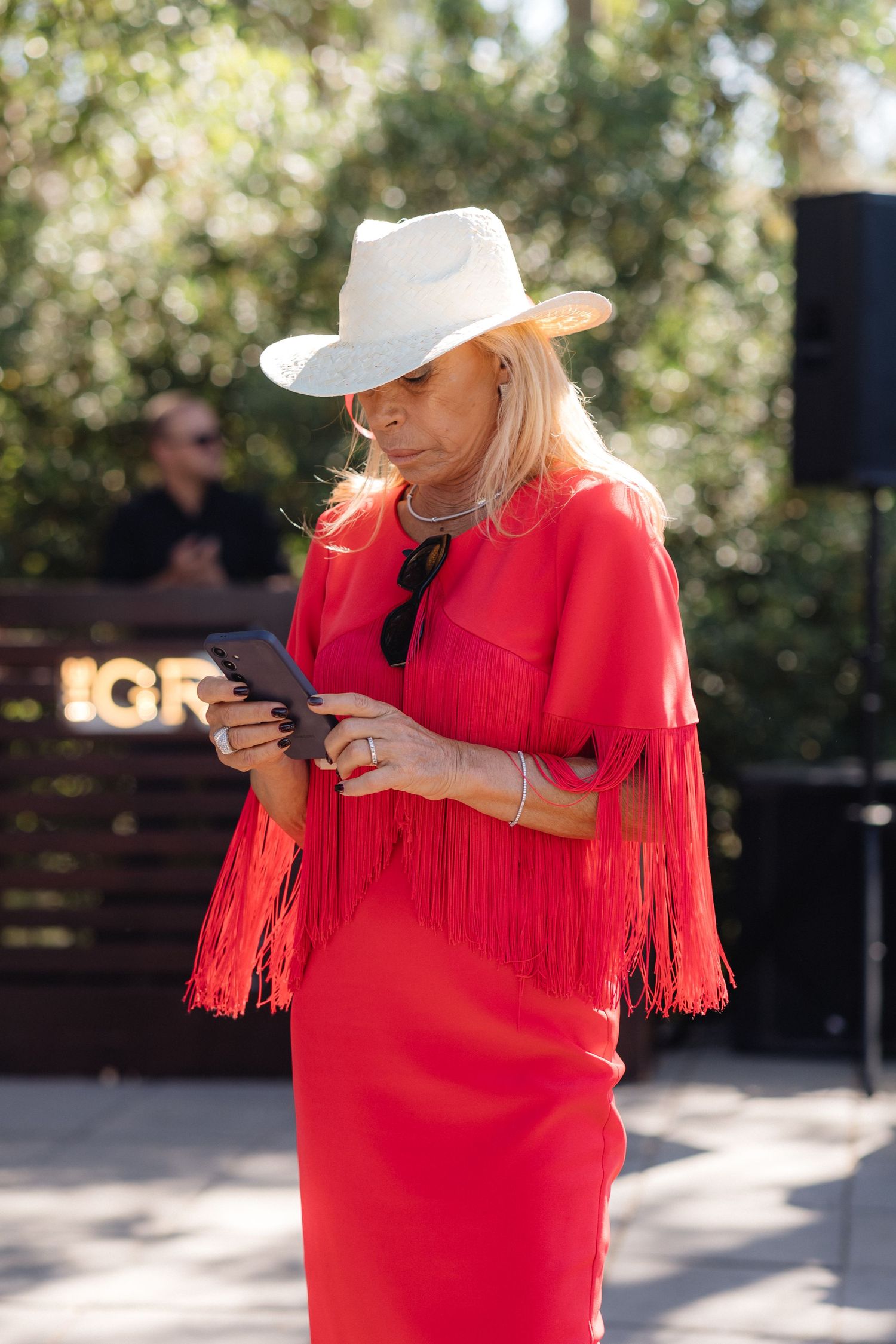 Person in bright red fringed dress and white hat looking at phone outdoors.