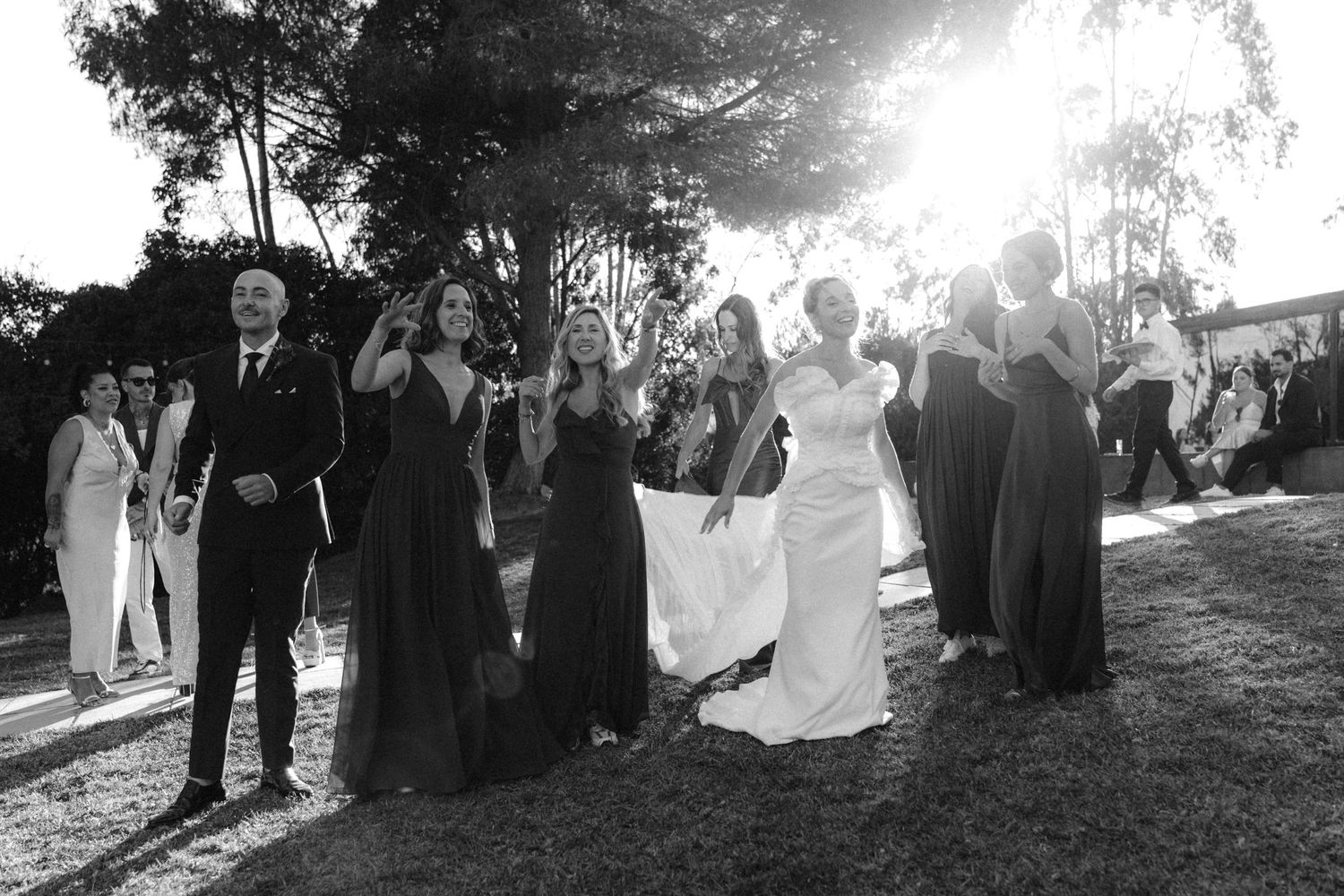 Wedding party poses outdoors in black and white photo with bridesmaids in dark dresses and sunlit background.