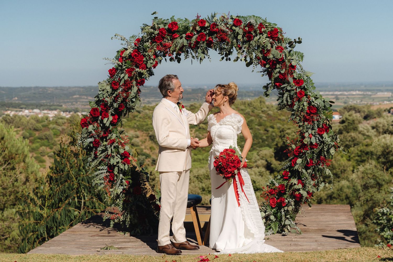 A couple stands under a stunning red floral wedding arch against a scenic landscape backdrop.