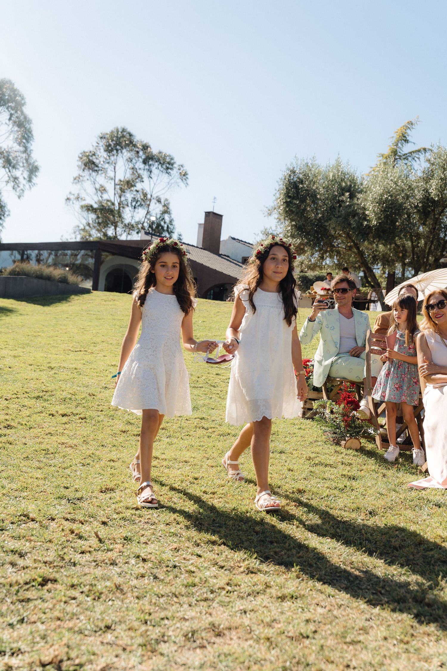 Wedding attendees in white dresses walk across a sunny hillside during an outdoor celebration.