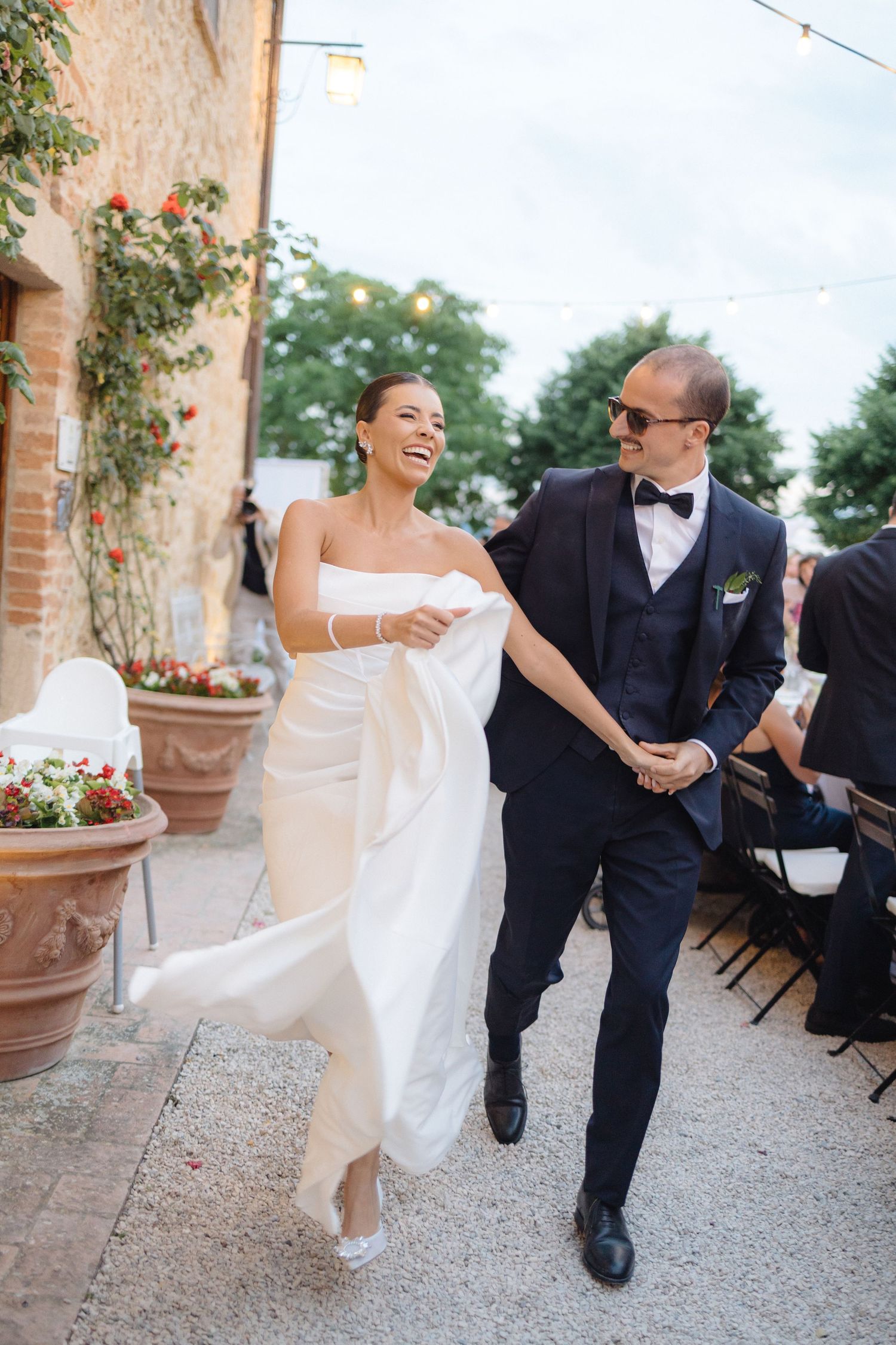 Laughing bride in strapless gown and groom in navy tuxedo entering outdoor wedding reception venue.