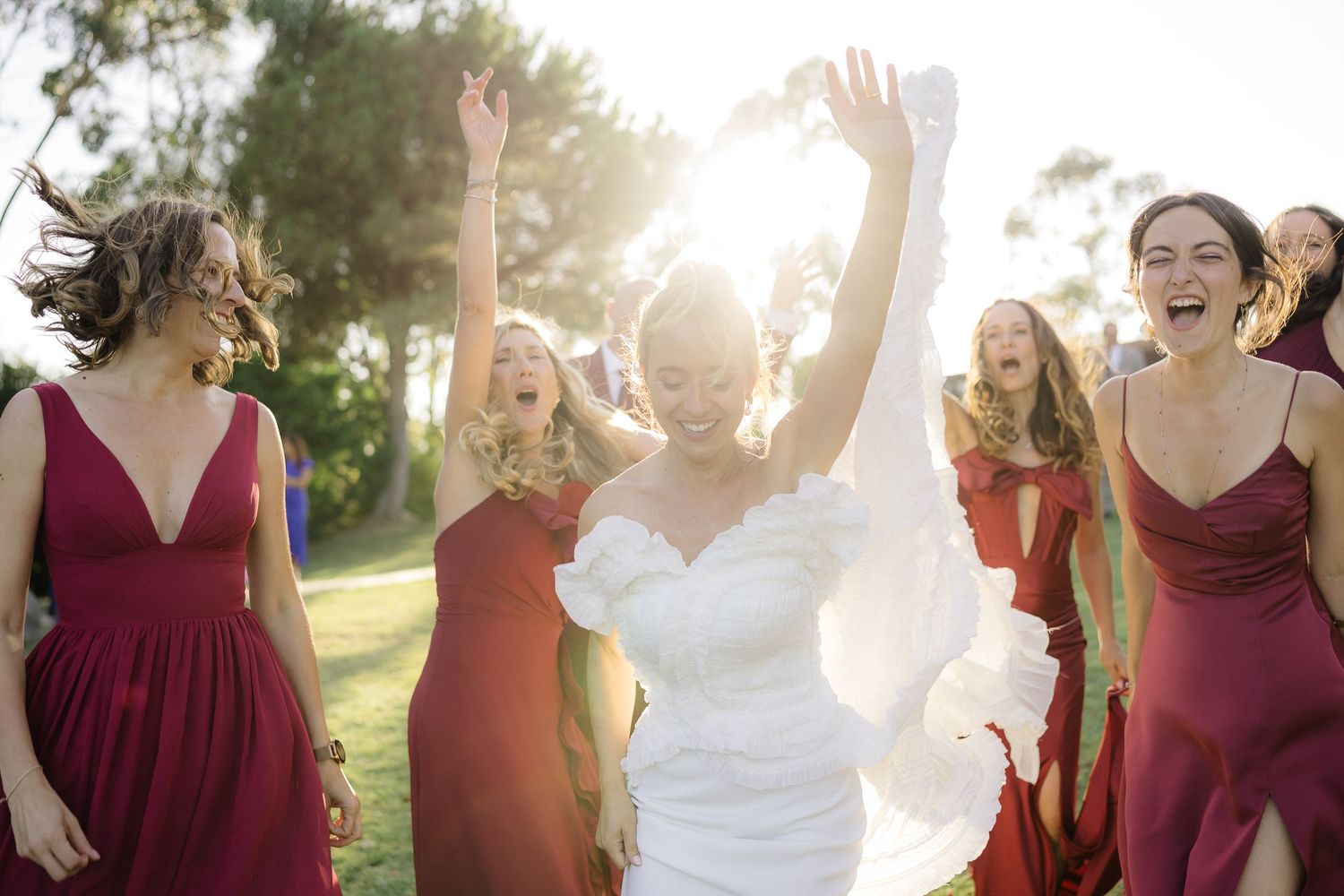 Wedding party in burgundy dresses celebrates joyfully in sunlit outdoor setting.