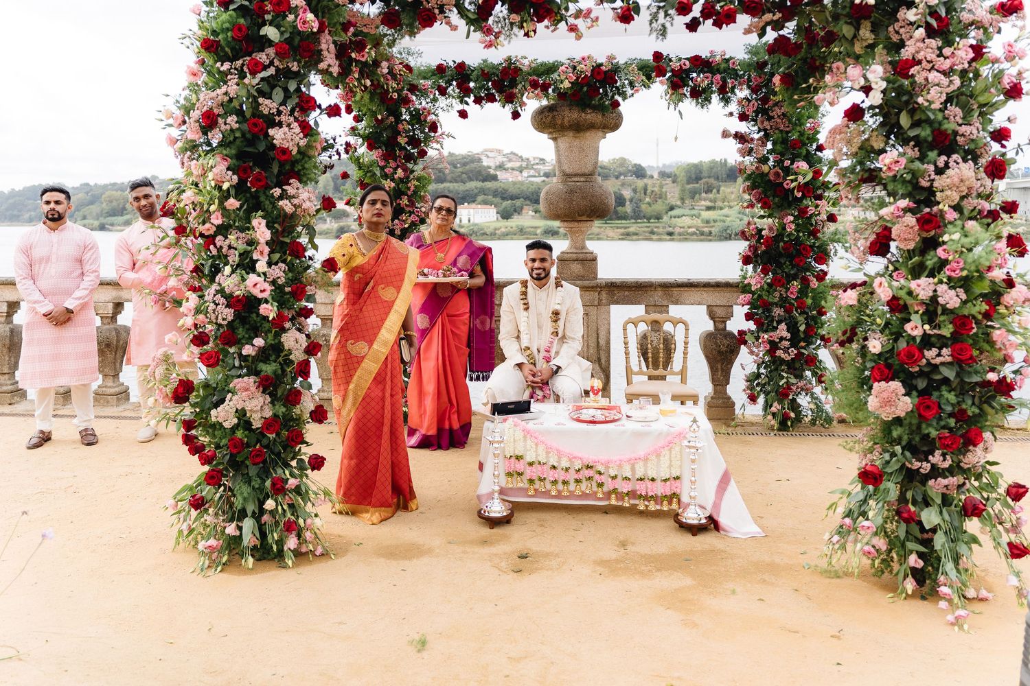 Wedding ceremony setup with red and pink floral arch against a waterfront backdrop.
