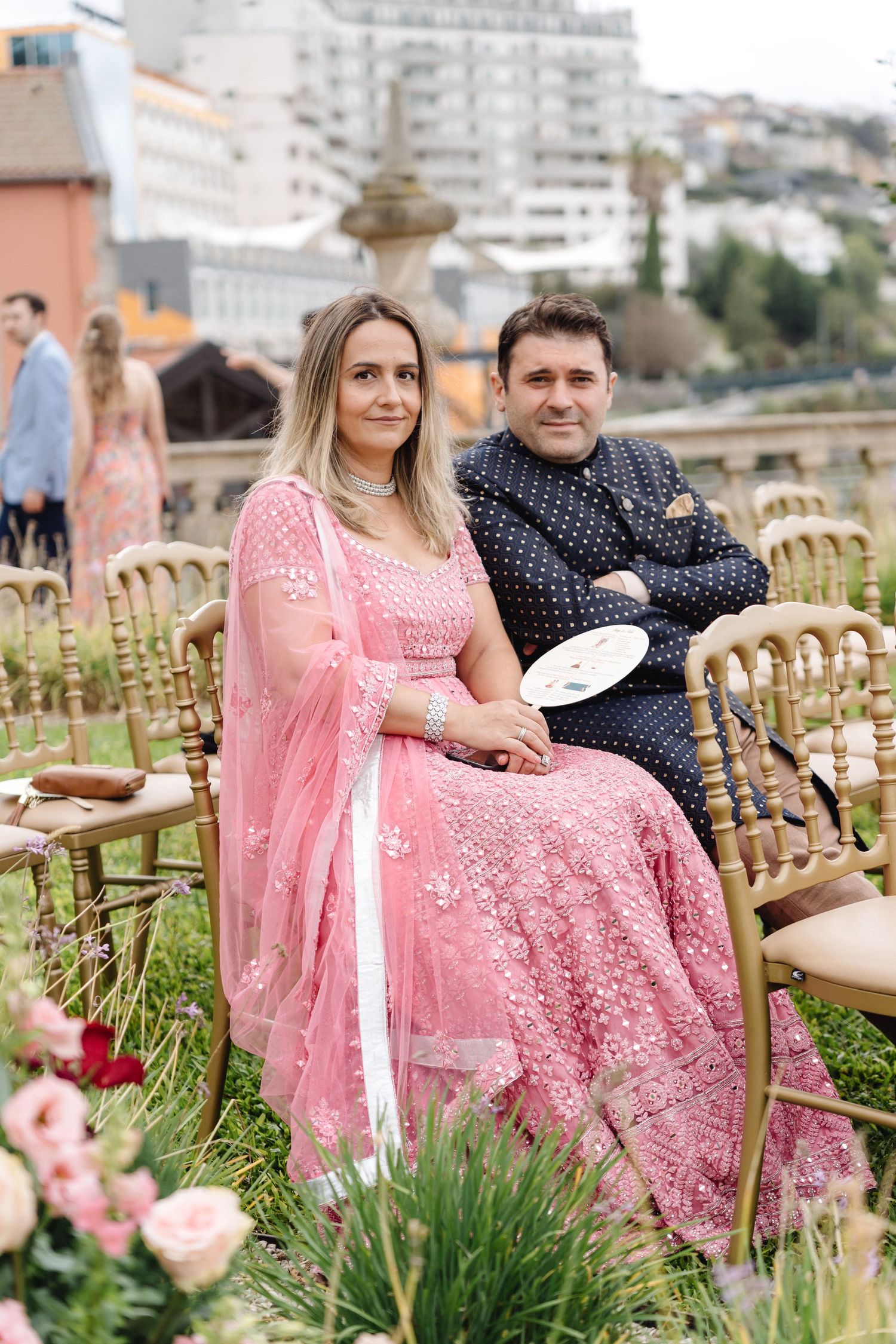Wedding guests seated in golden chairs among flowers with pink and navy formal attire.
