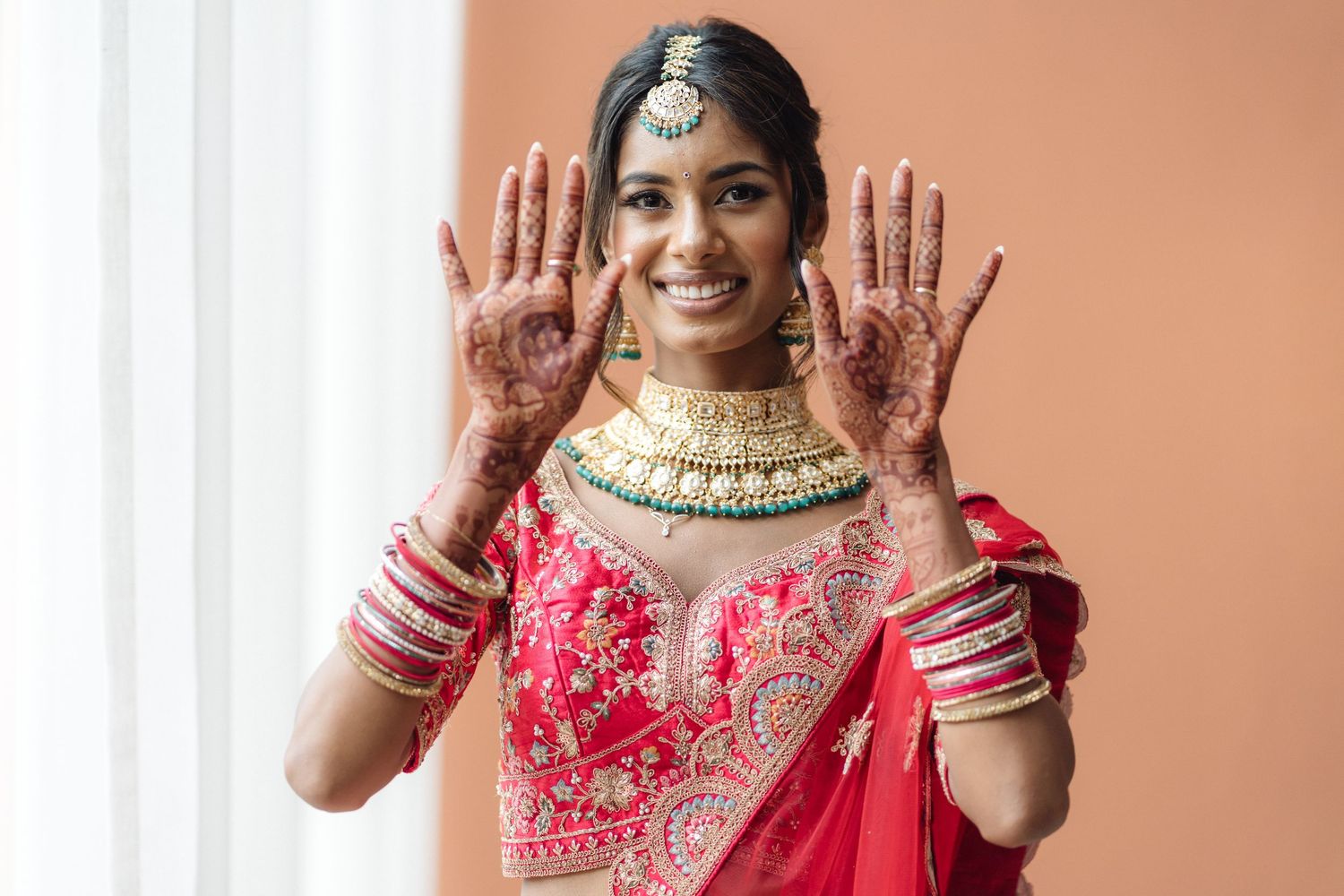 Bride in red traditional Indian attire showing decorated hands with henna and jewelry.