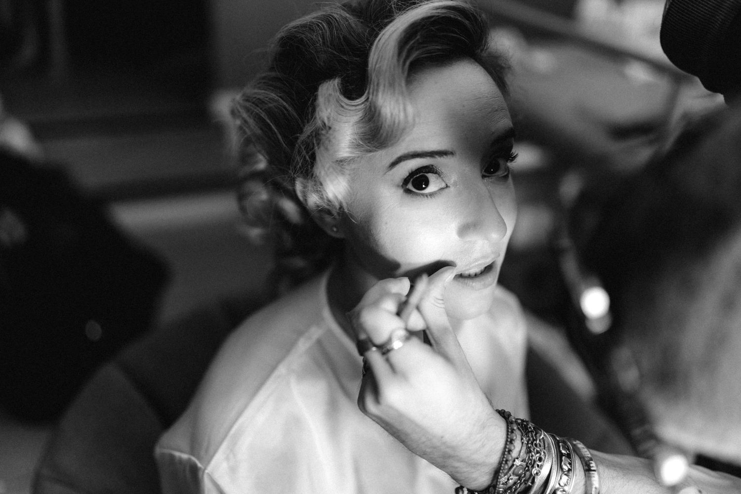 A black and white close-up portrait shows someone with curled hair wearing jewelry and biting their fingers nervously.