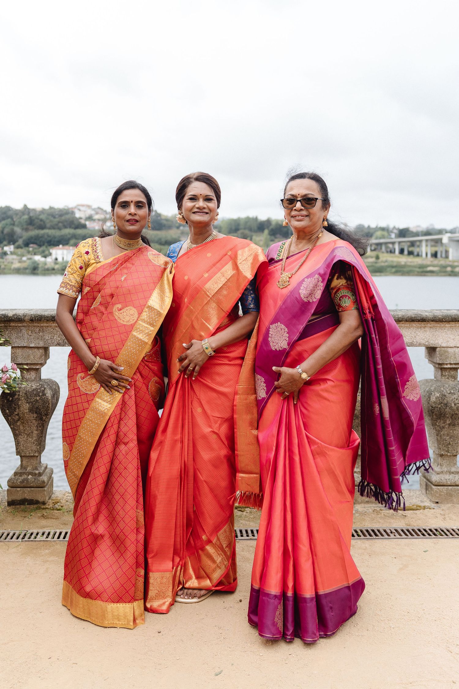 Group photo of women wearing vibrant silk saris in coral, red and magenta colors by a waterfront.