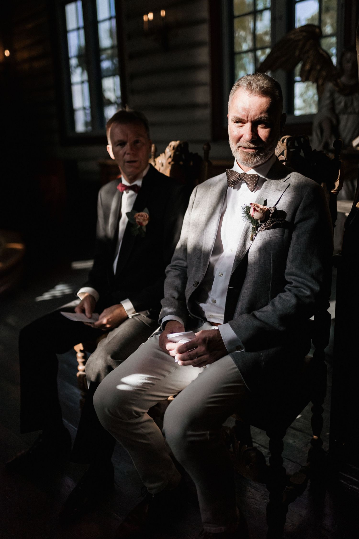 Men in formal suits sitting in dramatic lighting at wedding ceremony.