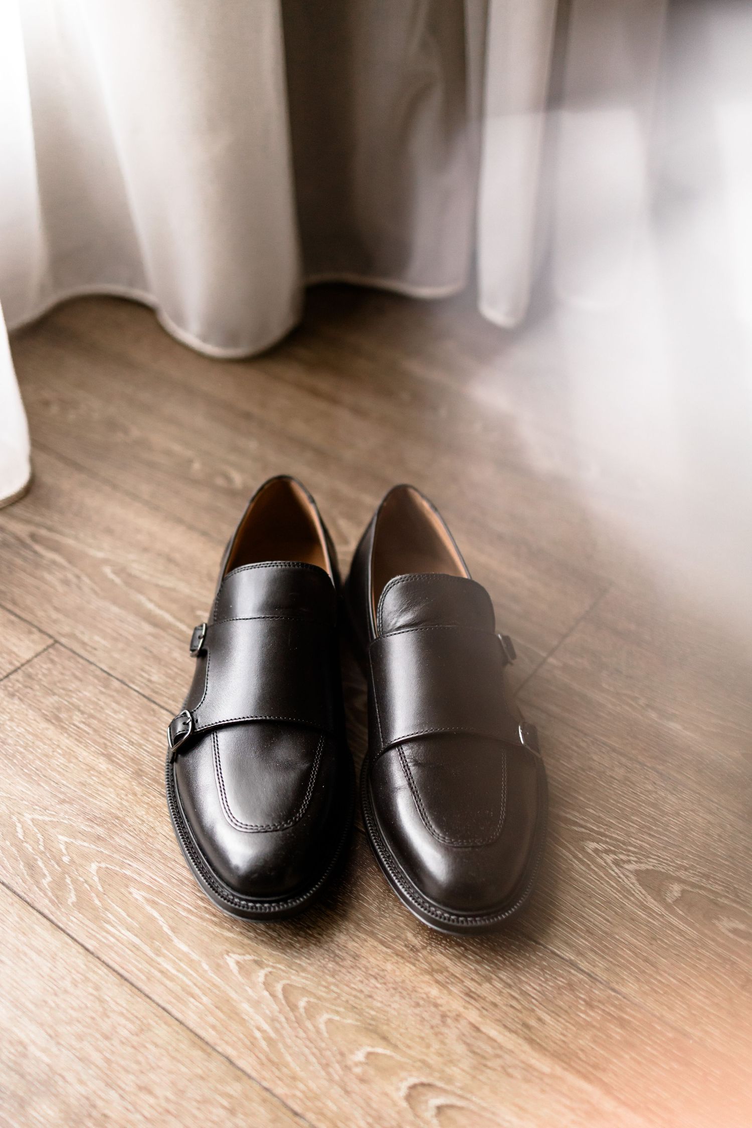 A pair of black leather dress shoes sits on a light wooden floor against white curtains.