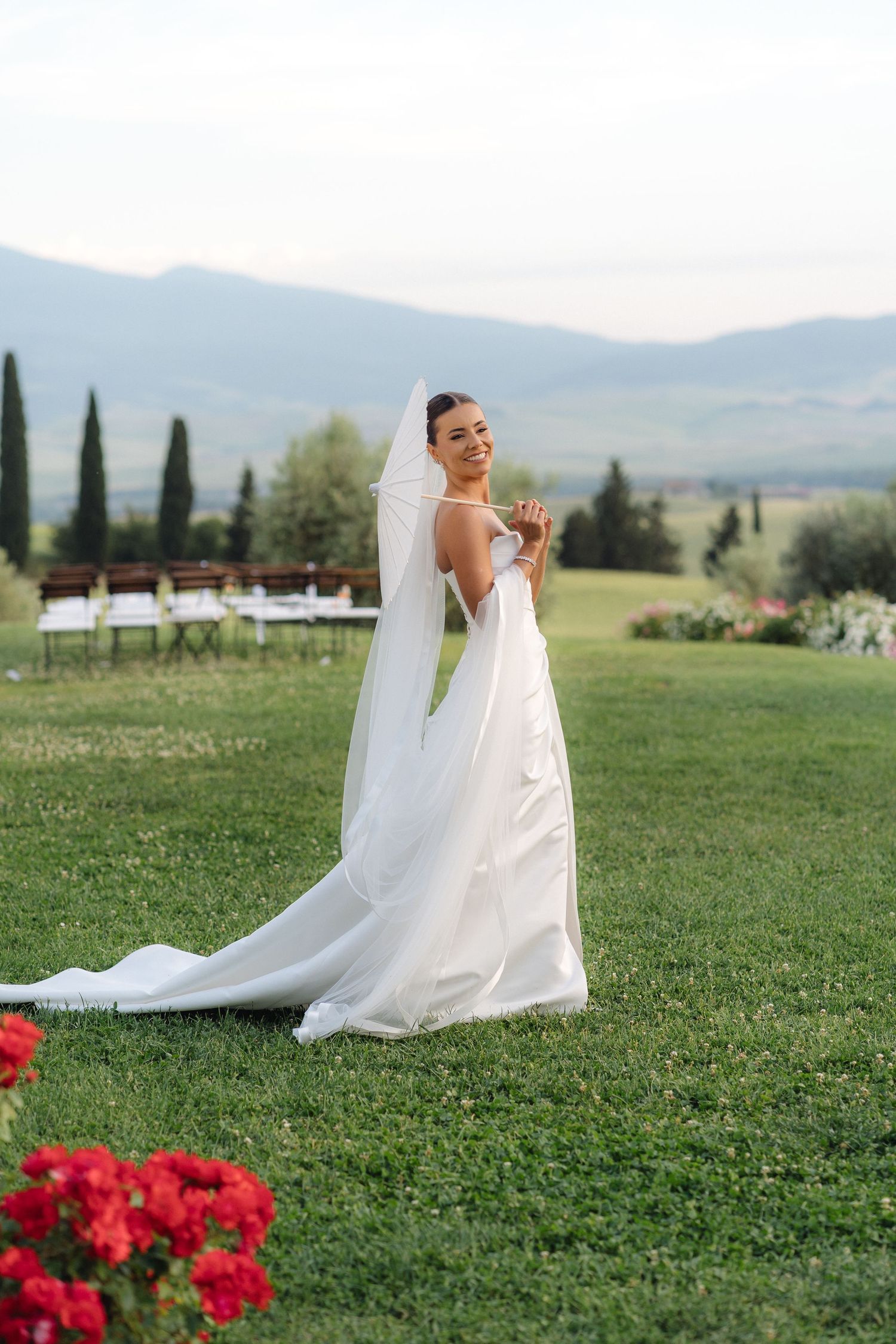 Smiling bride in a white gown and veil standing on a green lawn with mountain views at sunset.