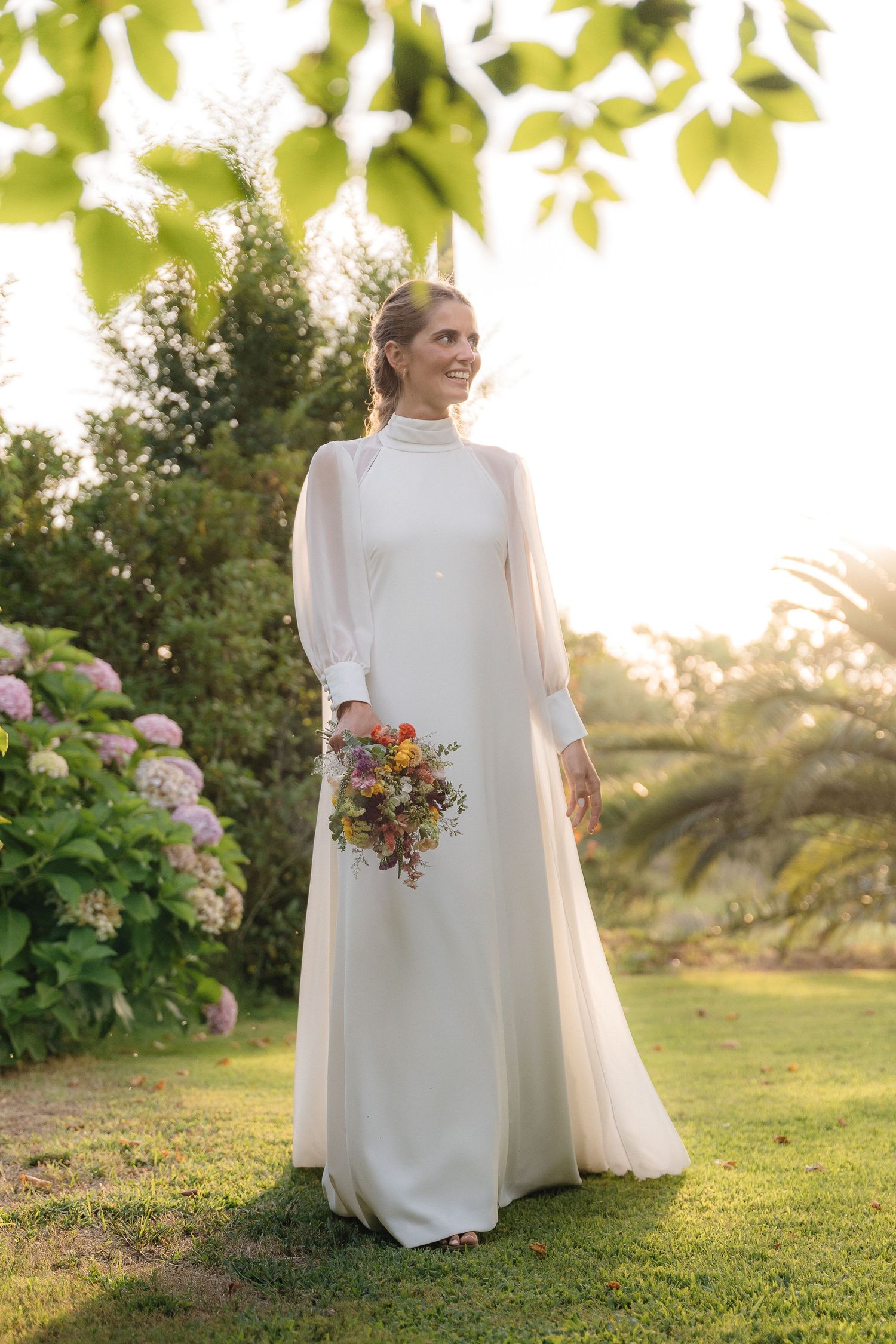 Elegant bride in minimalist white dress poses in garden setting with bouquet.