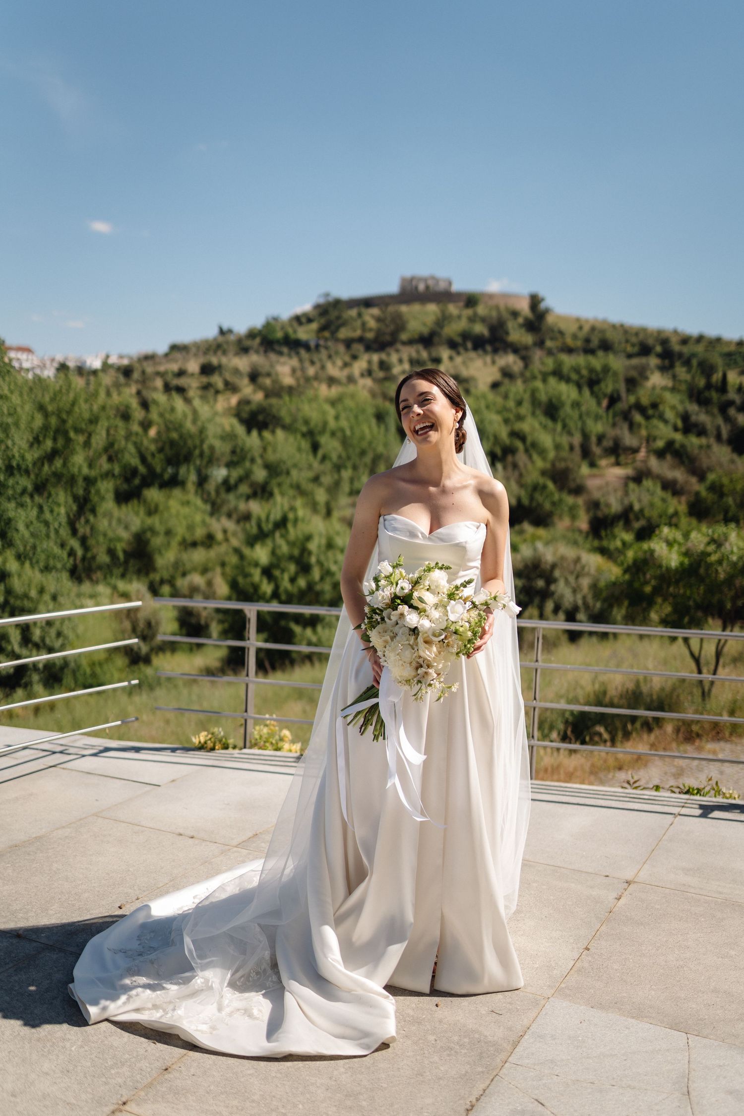 A bride in a flowing white dress poses with her bouquet against a scenic hillside backdrop.