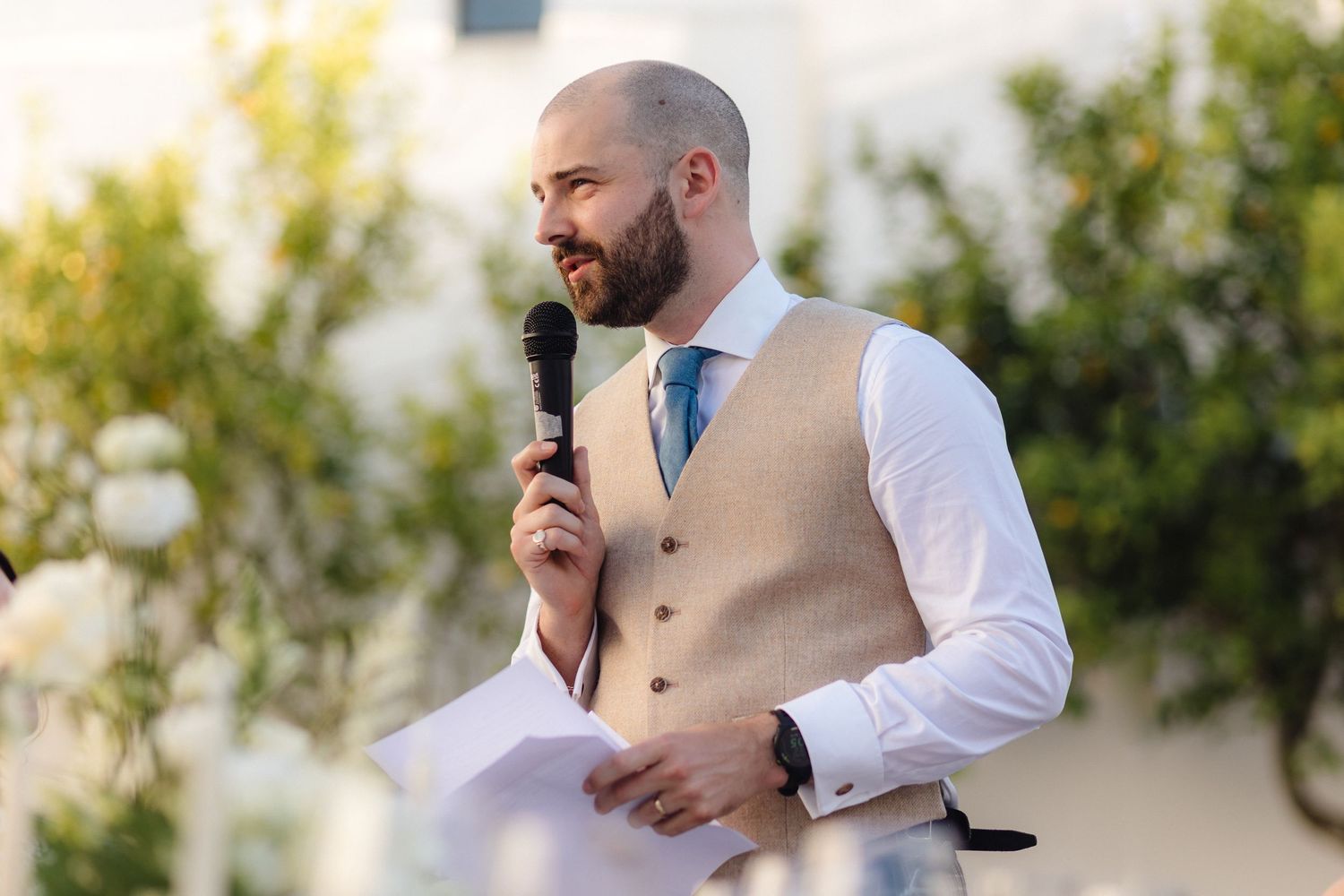 A speaker in a beige vest and blue tie delivers a speech at an outdoor wedding reception.