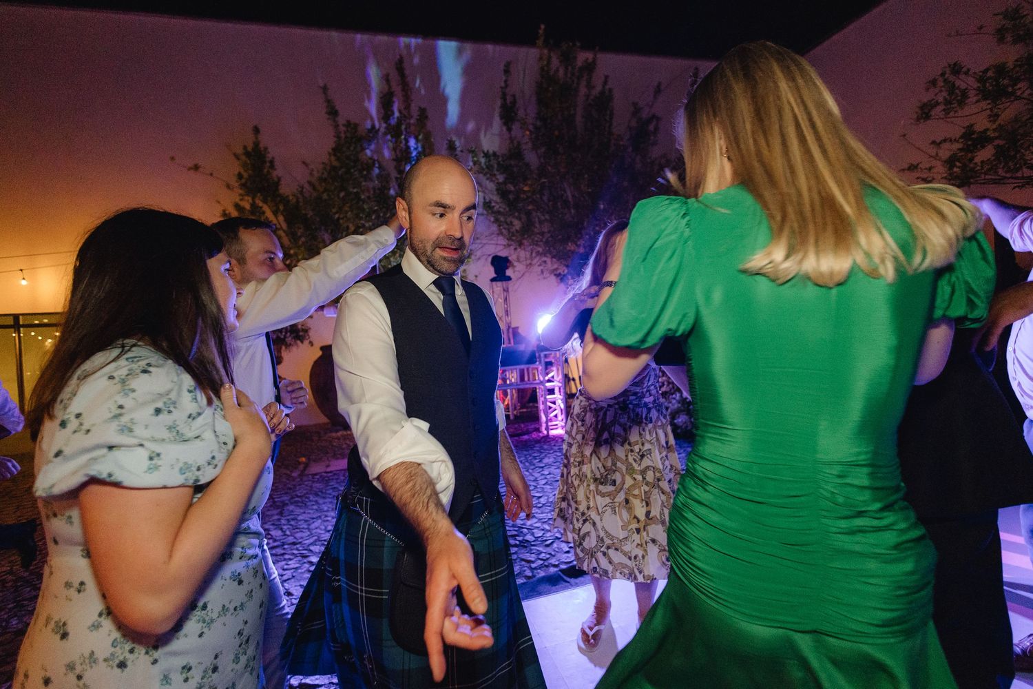 Wedding guests dance and celebrate on a dimly lit dance floor with colorful lighting effects.