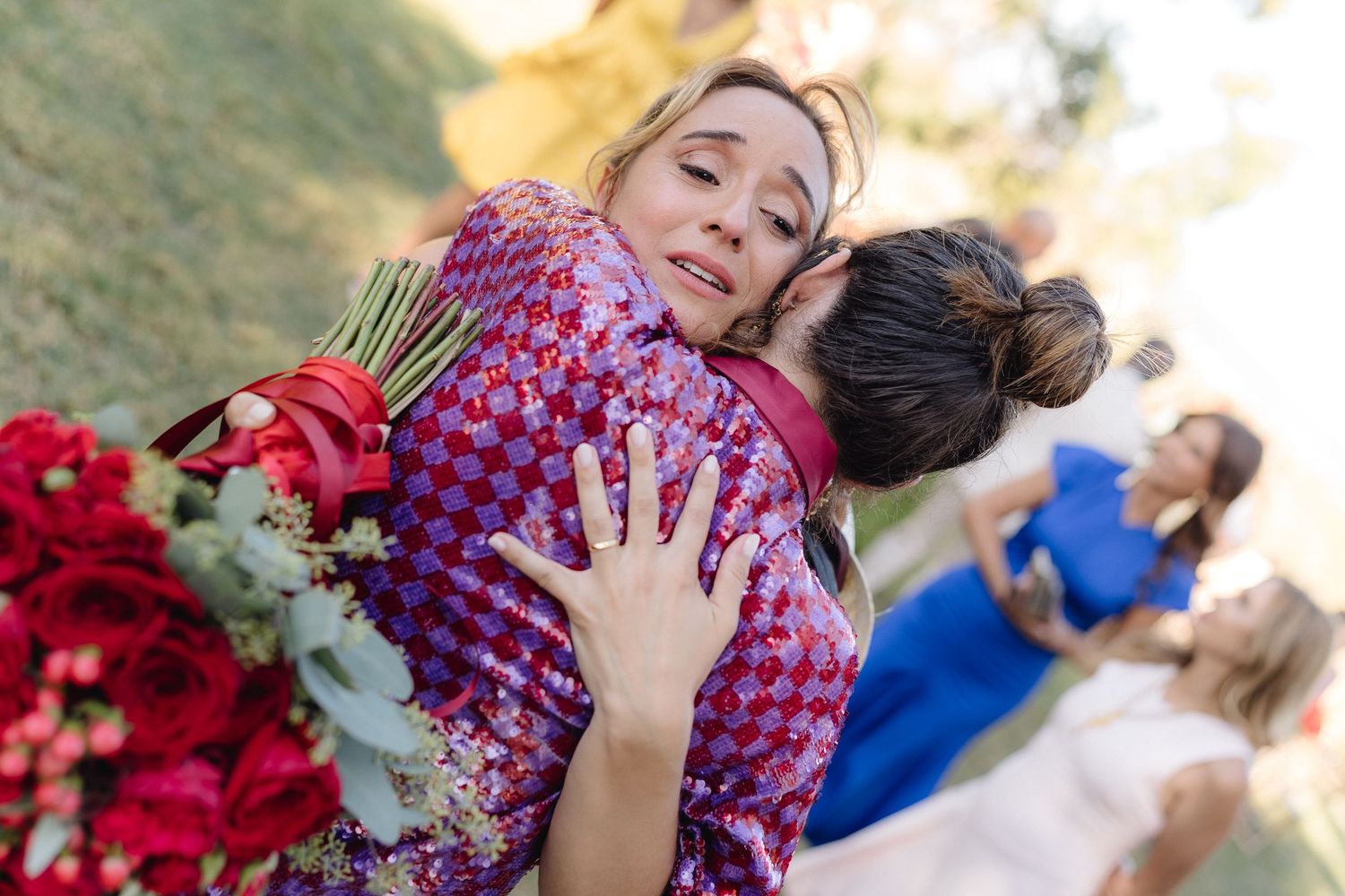 Two wedding guests share a joyful hug while holding a bouquet of red flowers.