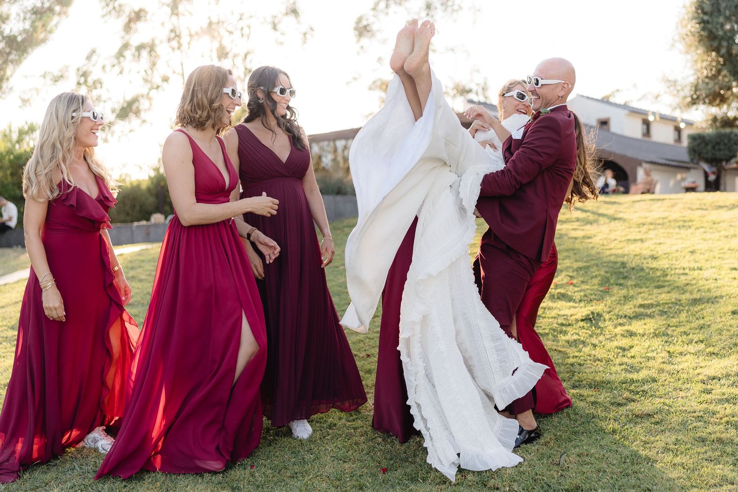 Wedding party in burgundy dresses dancing together on grass.