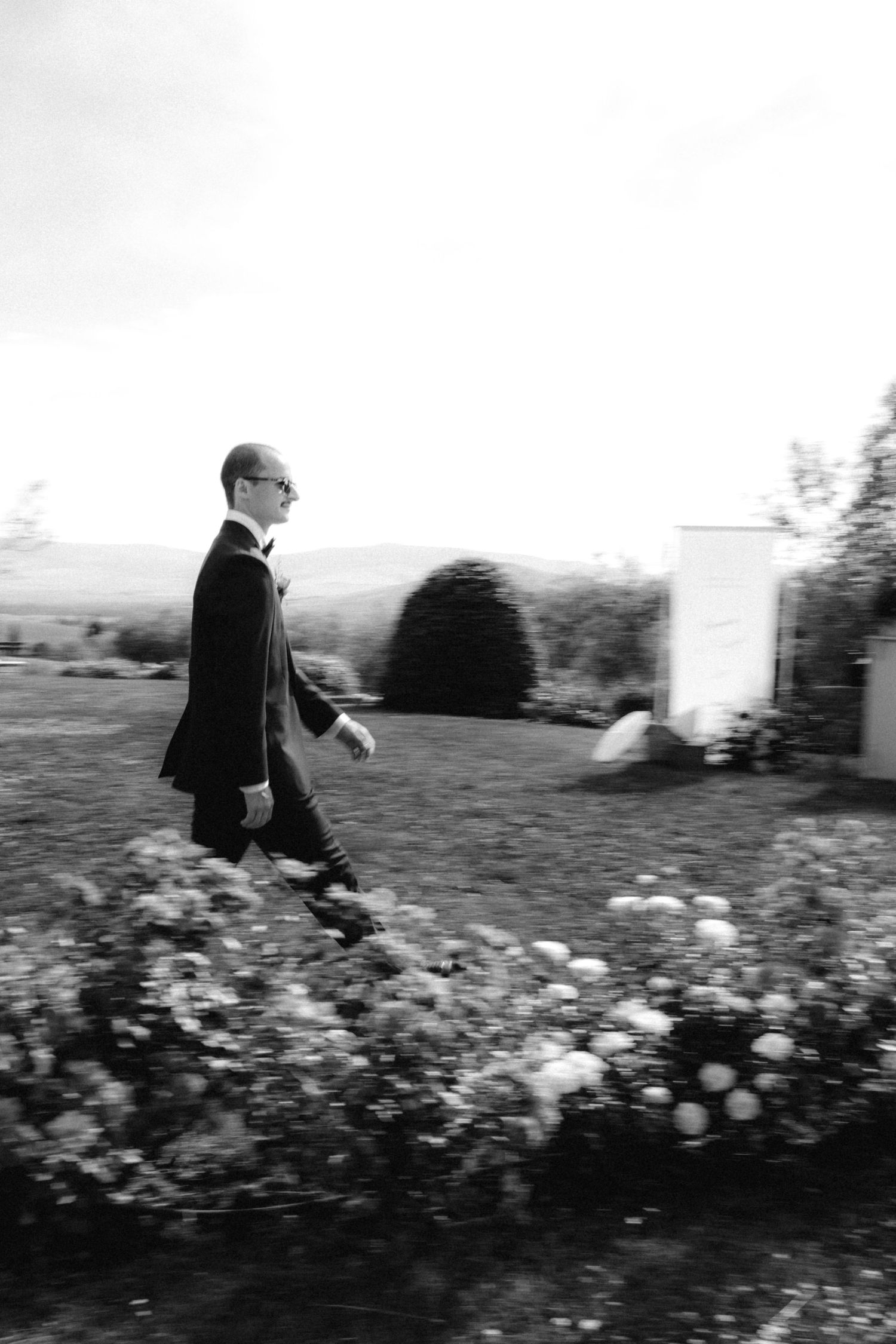 Groom in black tuxedo walking through garden with flowers in elegant black and white wedding photo.