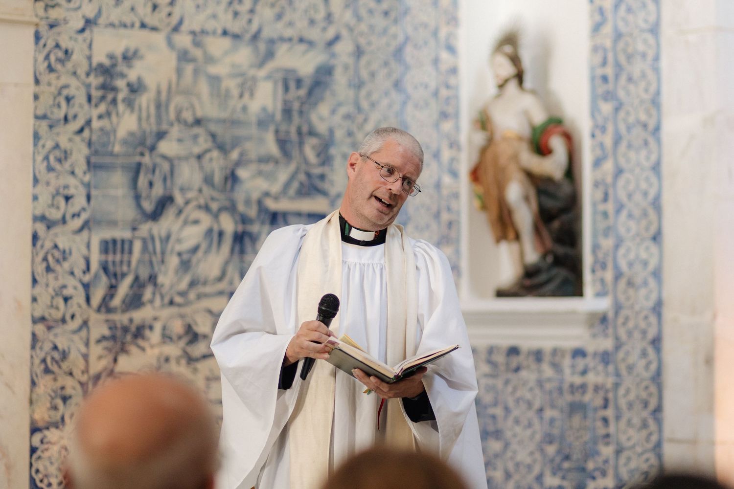 Priest in white robes reading from a book during a religious ceremony in front of blue and white tiled walls.