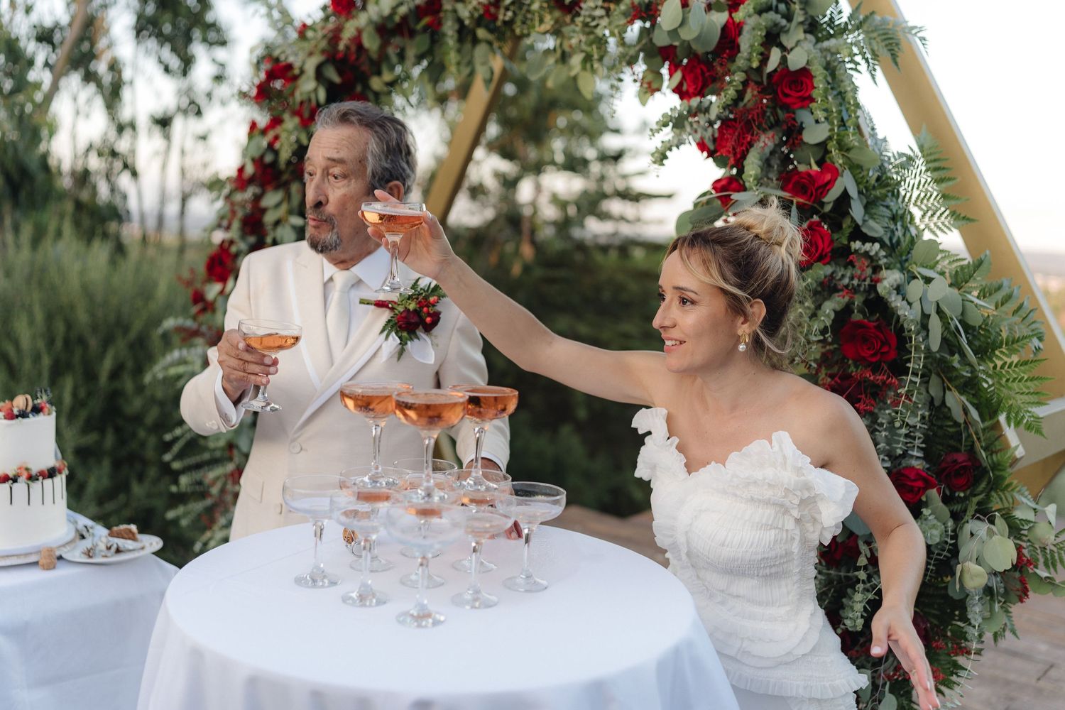 Wedding party pouring champagne into a tower of glasses outdoors with floral decorations.