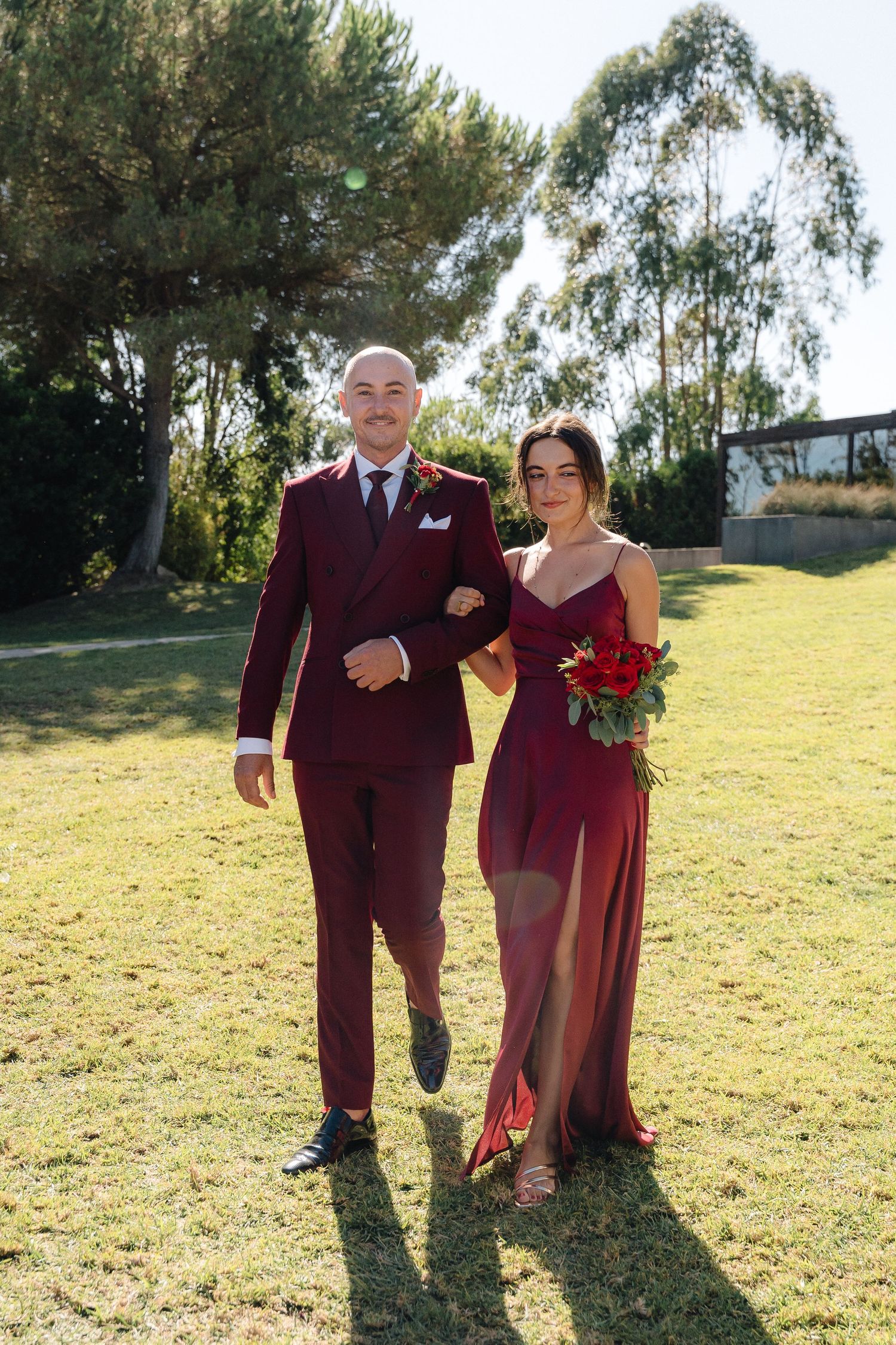 A couple in matching burgundy formal attire walks arm in arm on a sunny lawn.