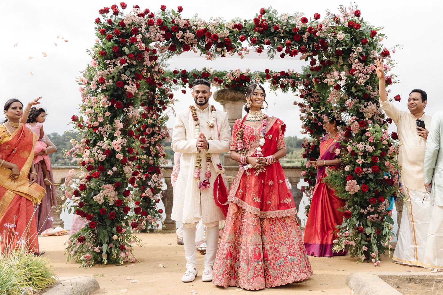 Traditional Indian wedding ceremony under a floral arch decorated with red roses and white flowers on a sunny day.