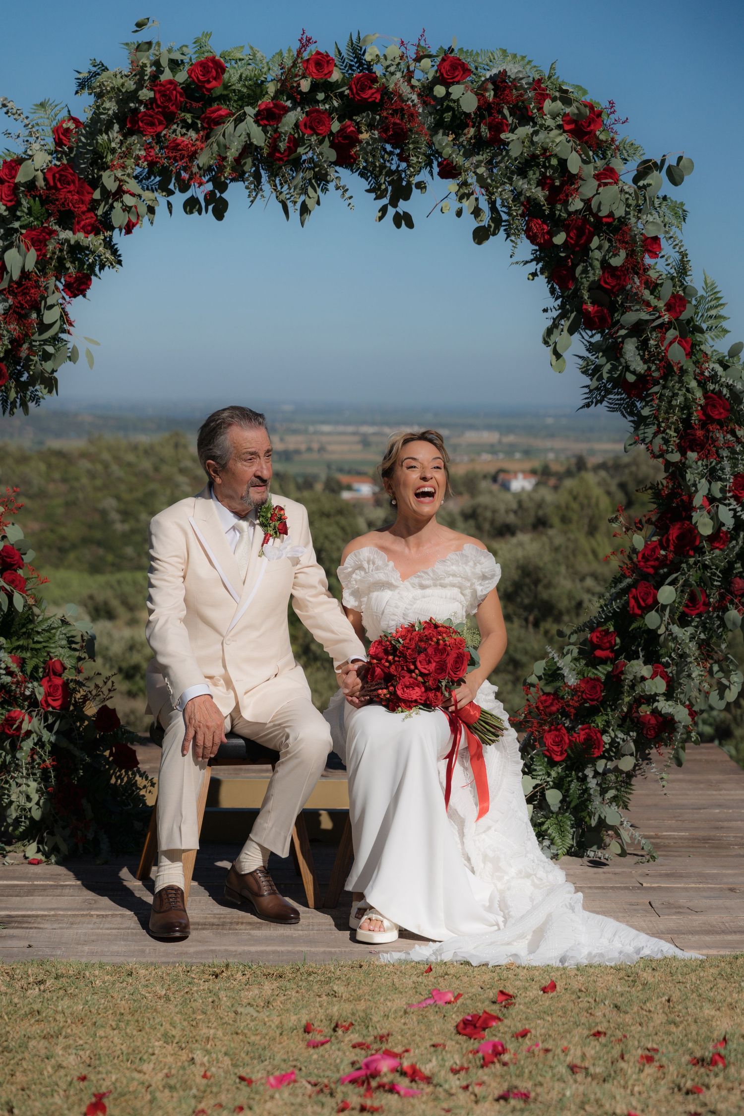 Newlyweds share joyful moments under a red rose floral arch in an outdoor garden setting.