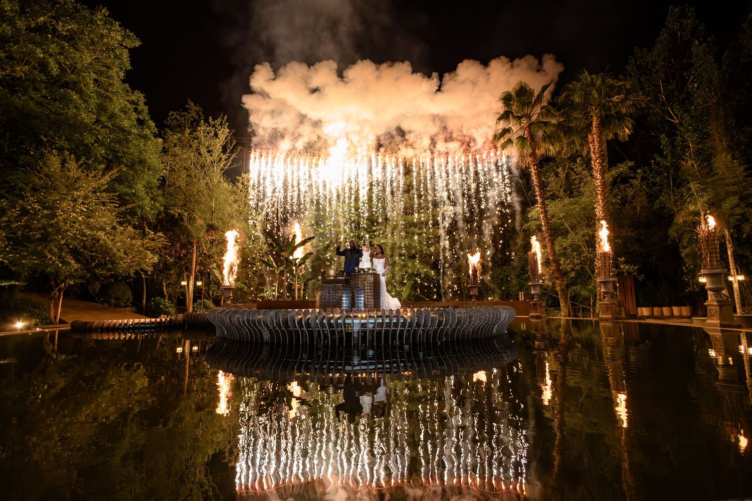 Spectacular fireworks display reflecting in a dark pond surrounded by trees.