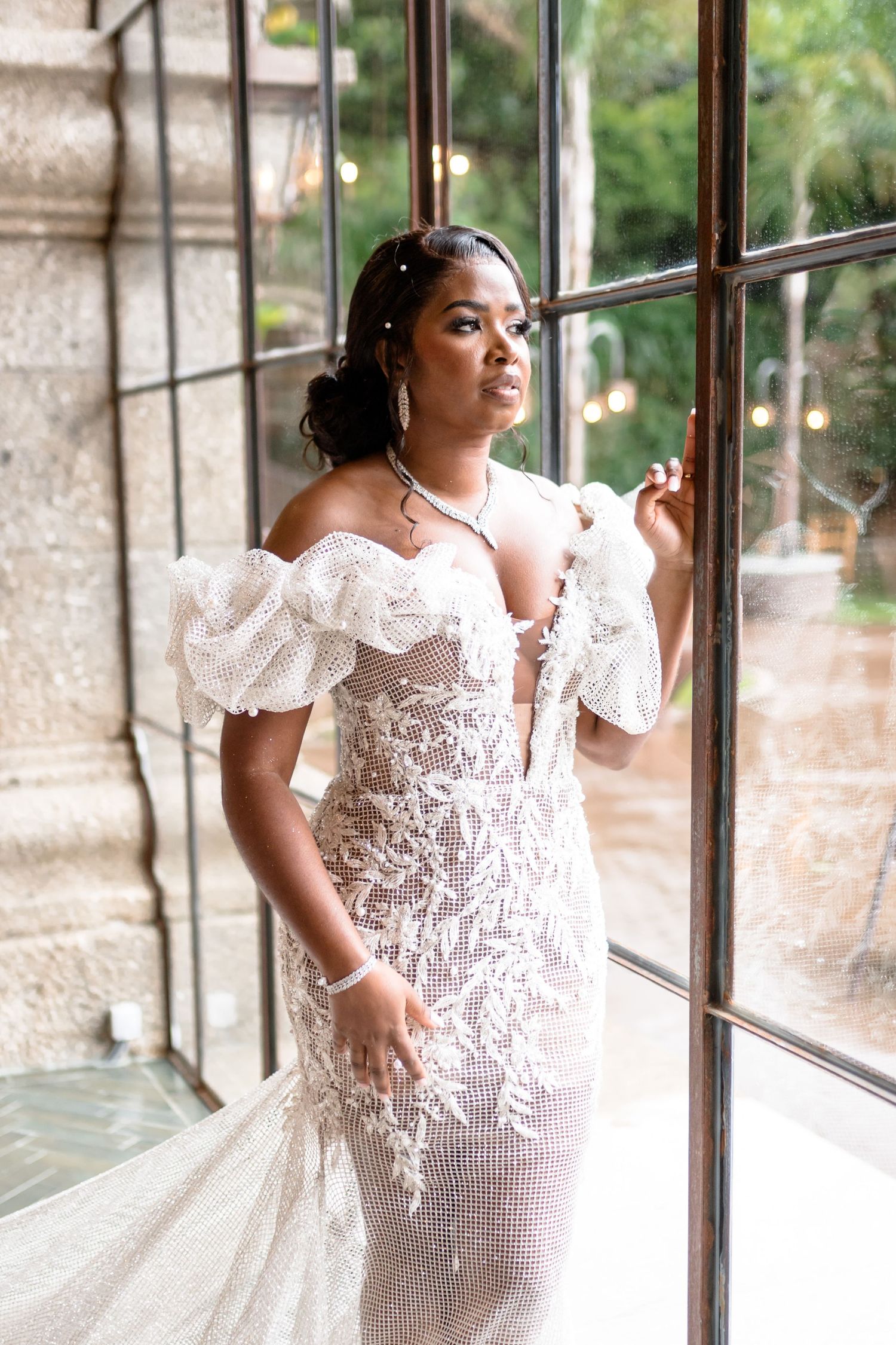 A bride in an elegant white lace dress poses by a window in natural light.