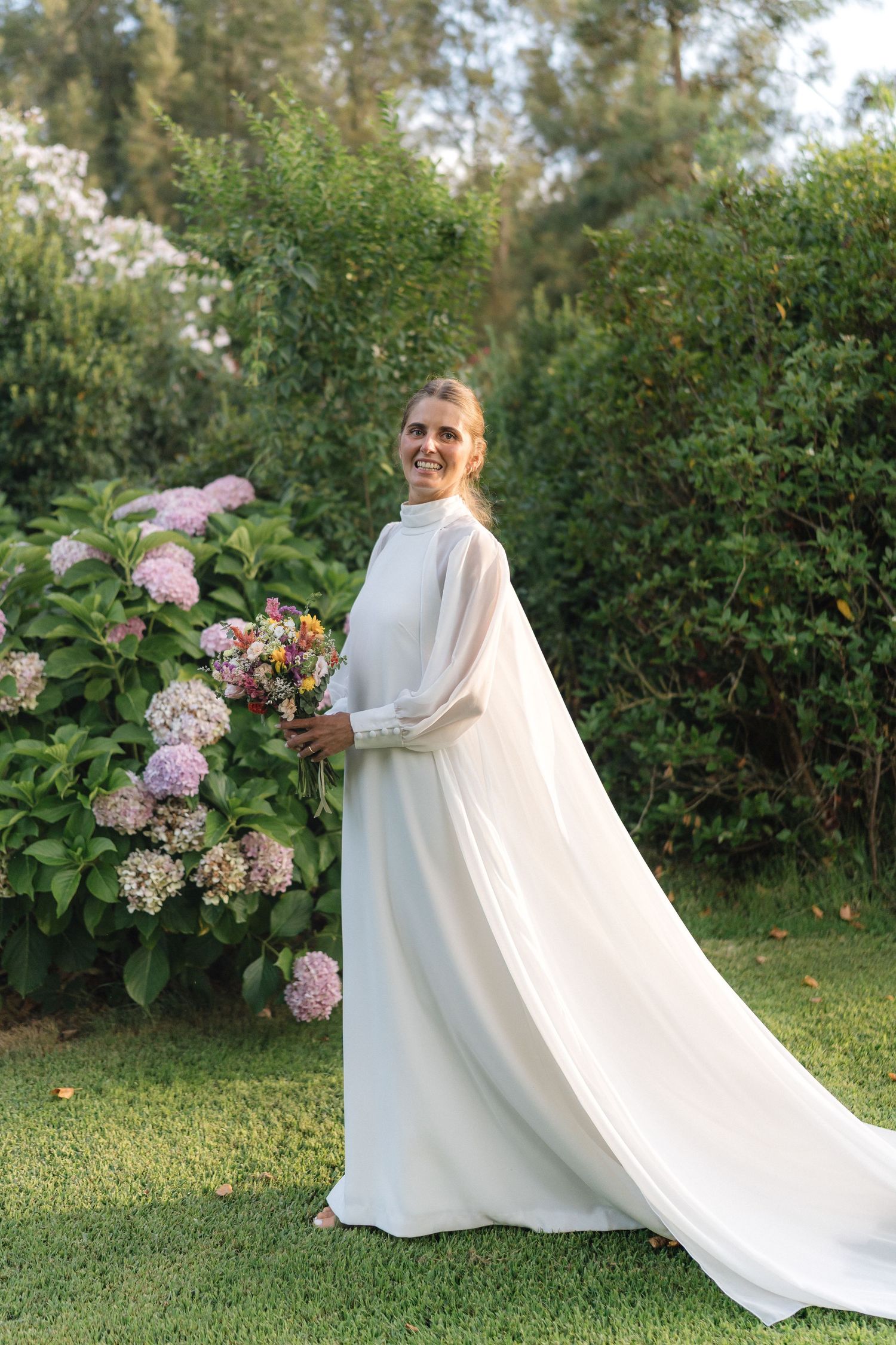 Elegant white wedding dress with flowing train against garden backdrop with pink hydrangeas.