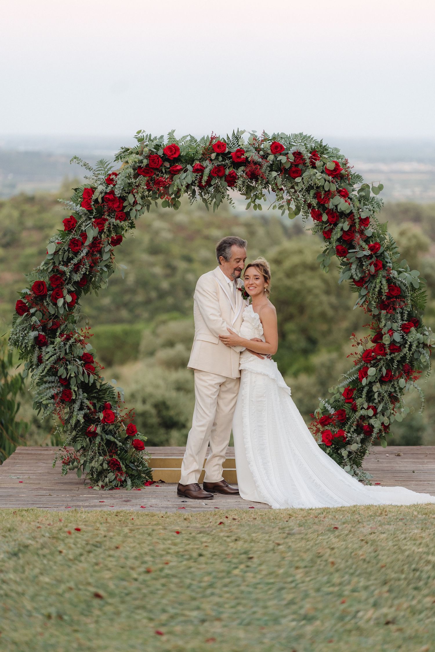 Wedding ceremony setting features beautiful red floral arch against scenic hilltop view.