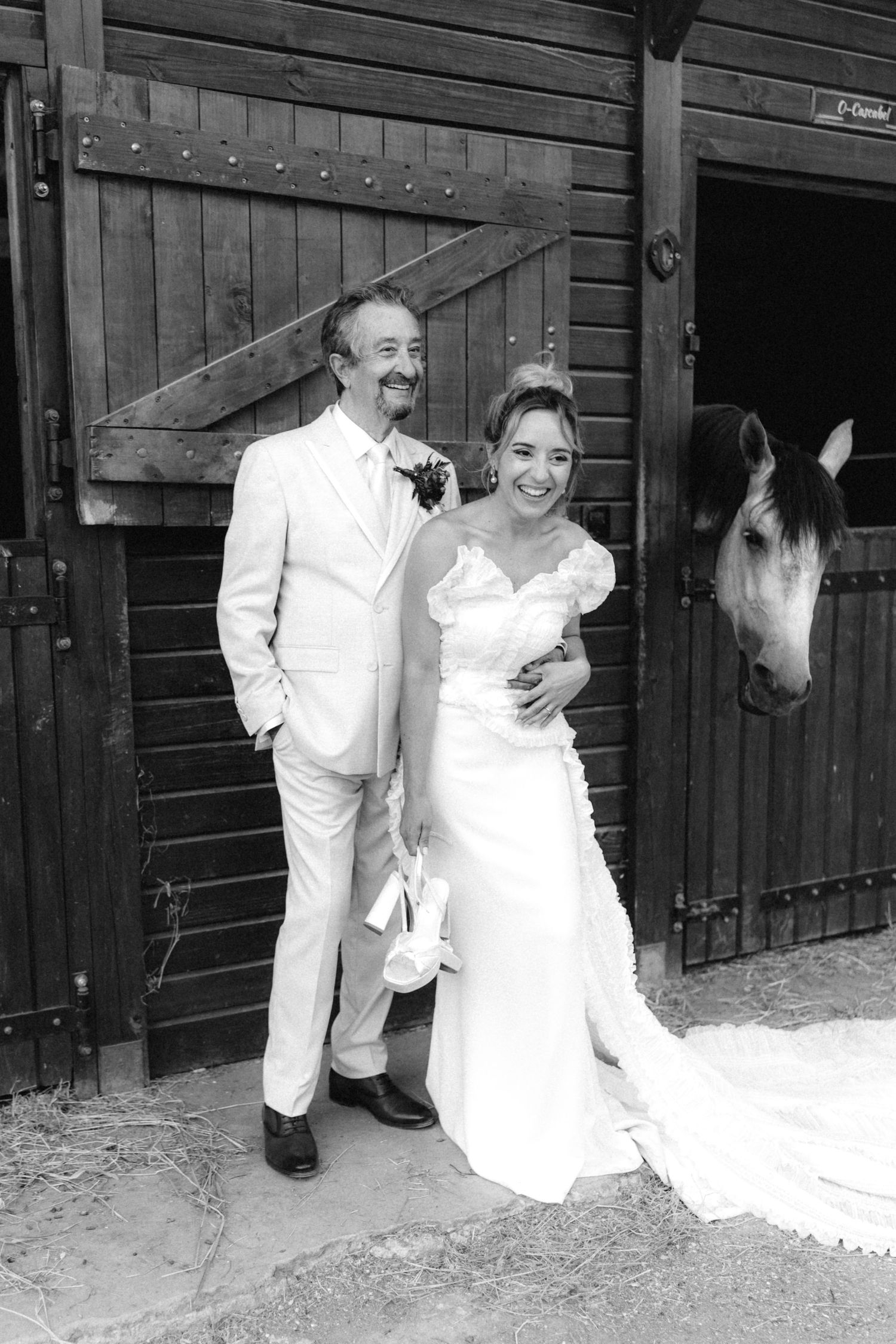 A couple poses with a white horse peeking out from a rustic stable door.